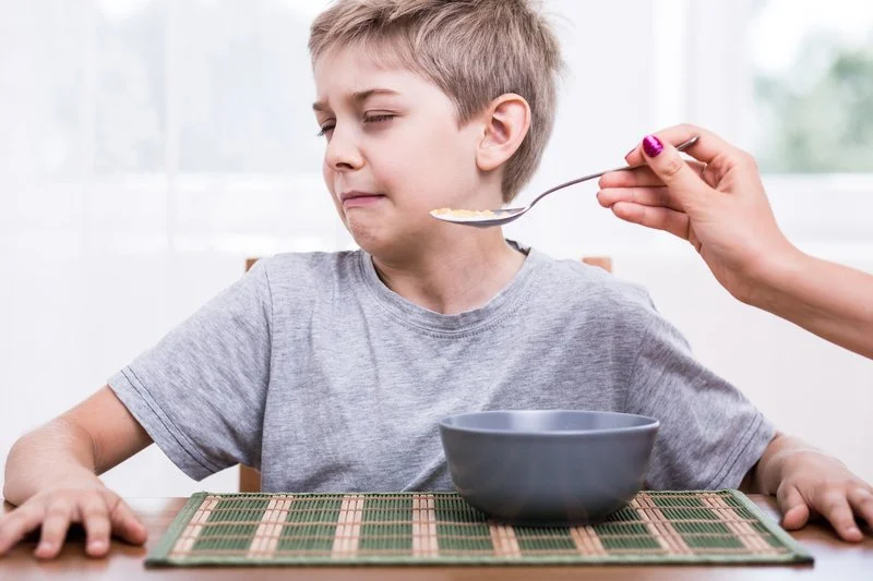 Child with a displeased facial expression sitting at a table with a gray bowl and a spoonful of food being offered, indoors with a blurred window background.