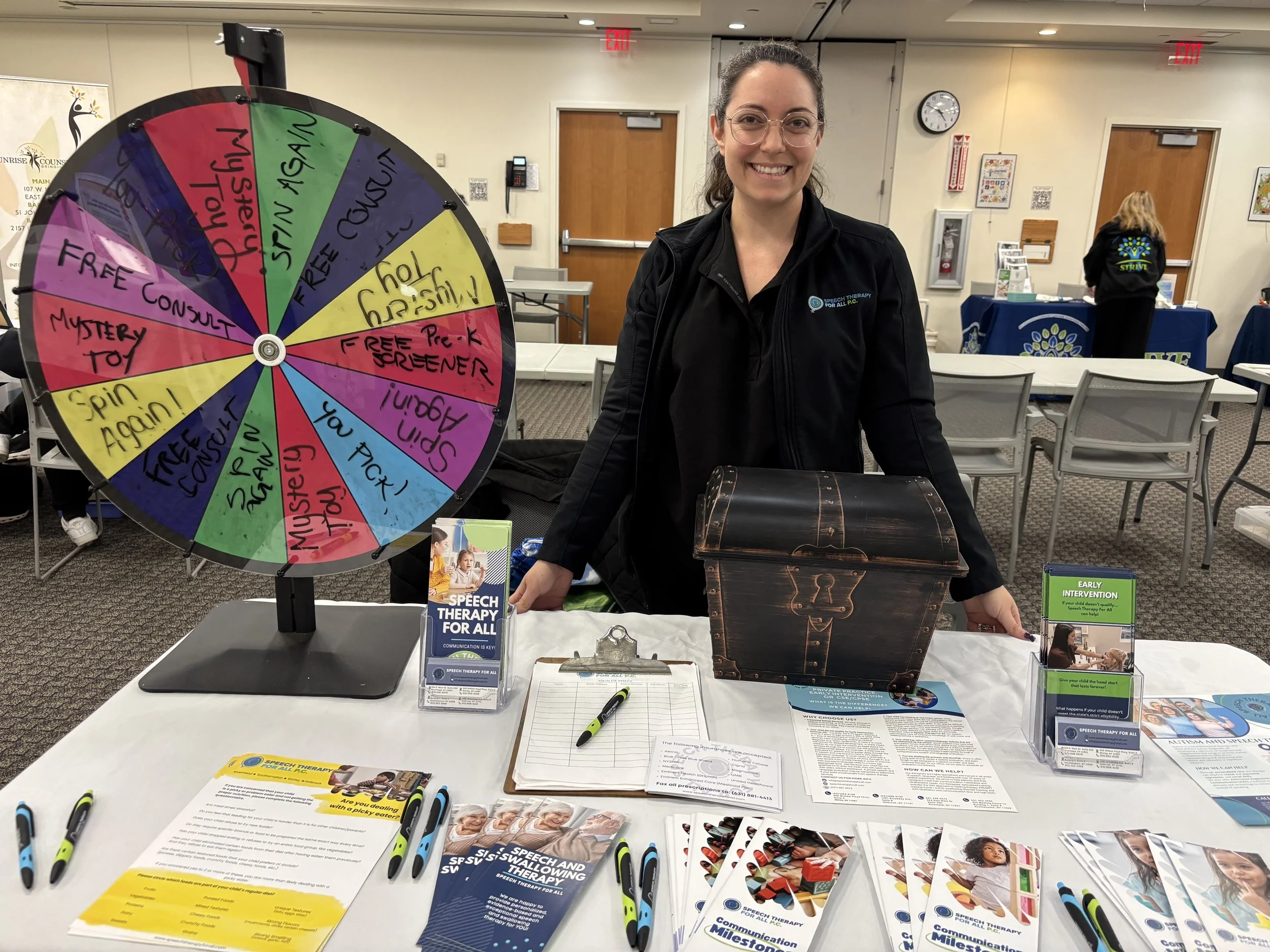A woman is standing behind a booth with a colorful spinning wheel that has various prize sections. The booth displays brochures and informational material about speech therapy. The woman is smiling and wearing glasses and a black jacket. In the background, there are other booths and chairs in a conference or community center setting.