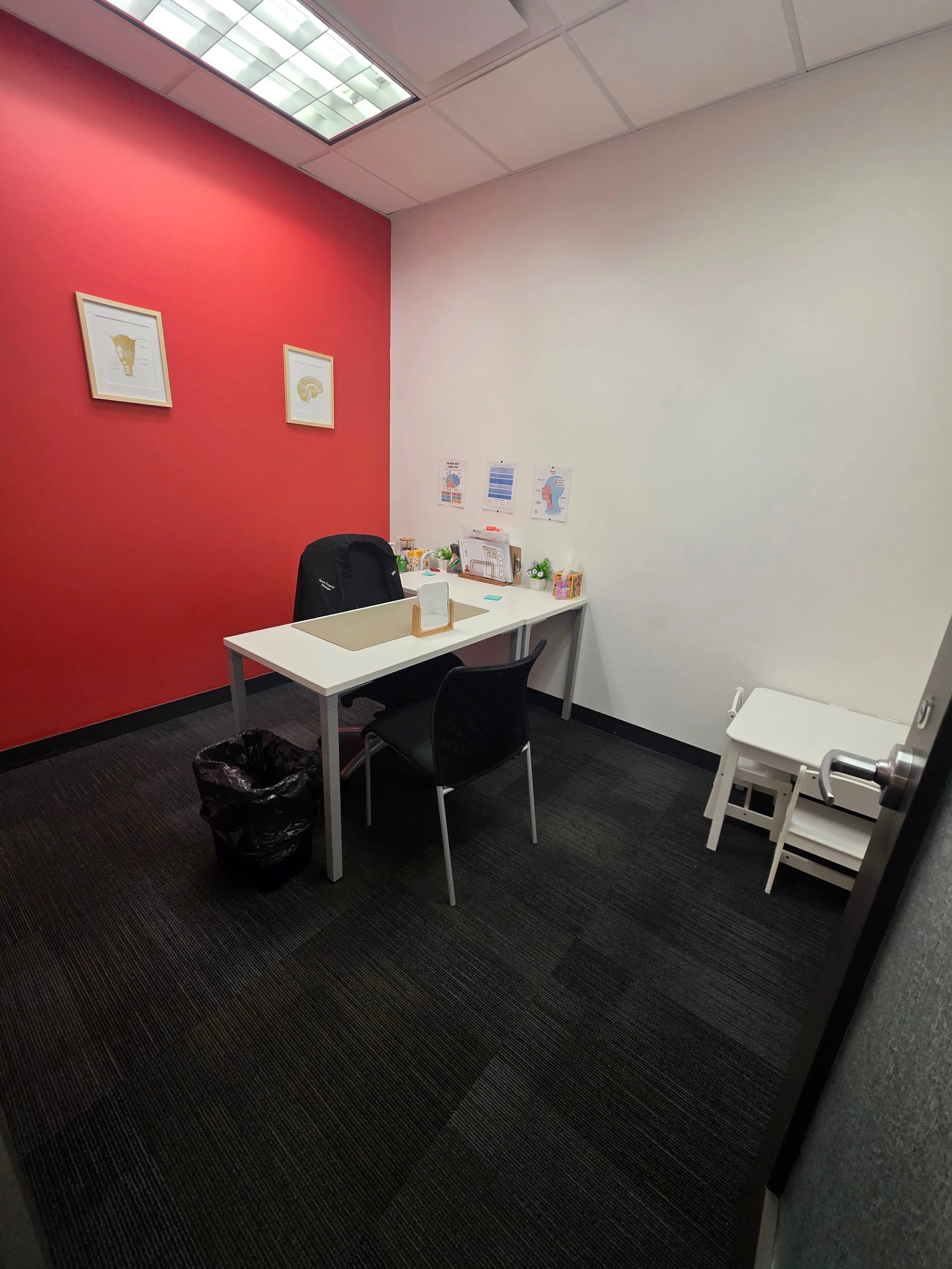 A small office with a white desk and black office chair. The desk has a black backpack, papers, and small decorations. The red accent wall has framed art, and the plain white wall has some papers with charts and a diagram. There is a small white tabl
