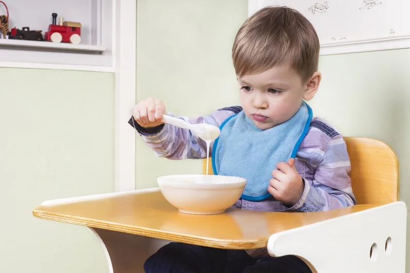 Young boy sitting in high chair, eating from a white bowl with a spoon in a kitchen or dining area.