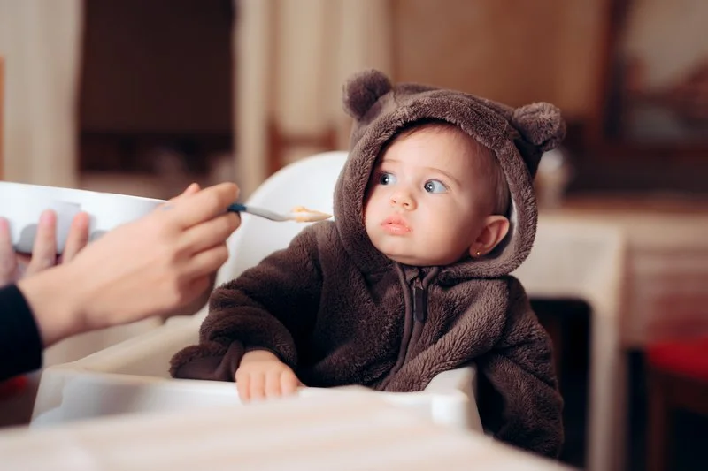 Baby dressed in a bear costume sitting in a high chair with a spoon and bowl, looking at the food.