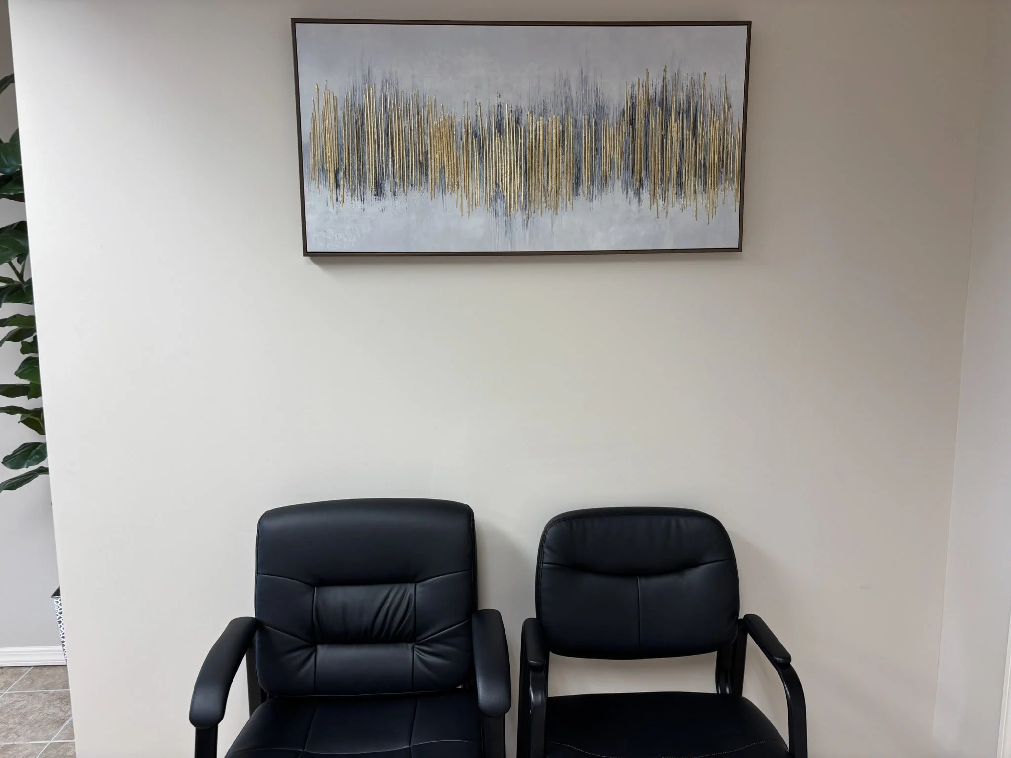 Two black leather office chairs in front of a beige wall with a framed abstract painting above them, and a part of a green plant on the left side.