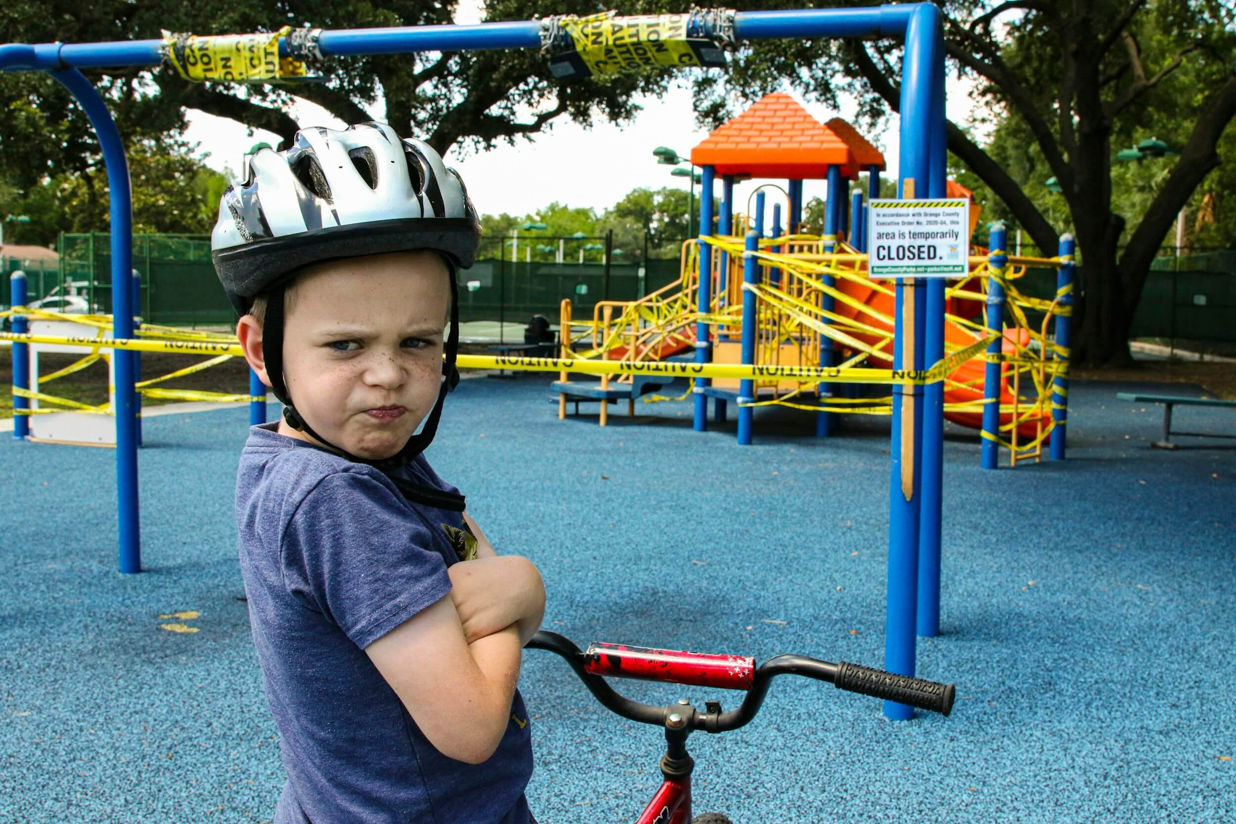 Young boy with a helmet on his head standing in a closed playground with a red bicycle, yellow caution tape, and a blue rubberized surface.