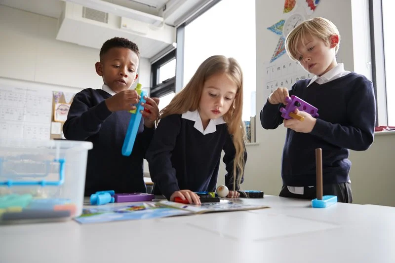 Three children in school uniforms playing with science toys at a table in a classroom.