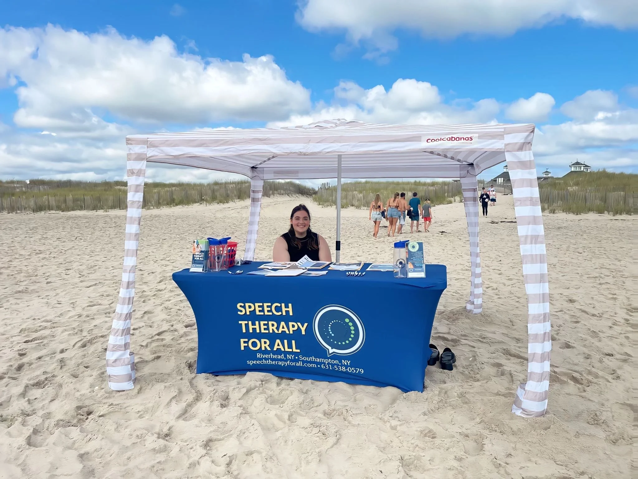 A woman sitting at a booth under a striped canopy on a sandy beach. The booth promotes speech therapy for all. There are brochures and supplies on the table, and a group of people walking in the background against a blue sky with scattered clouds.