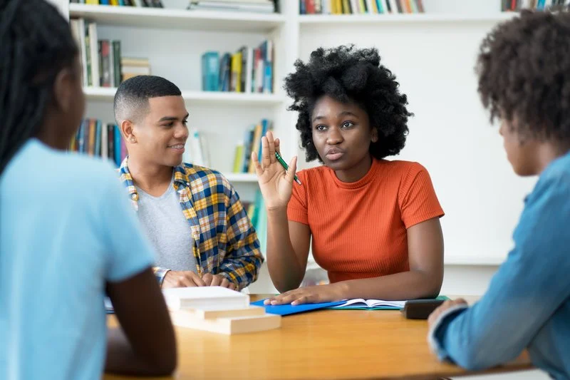 Four young people engaged in discussion at a table in a room with a bookshelf in the background. The woman in the center holds a green pen and appears to be speaking or explaining something.