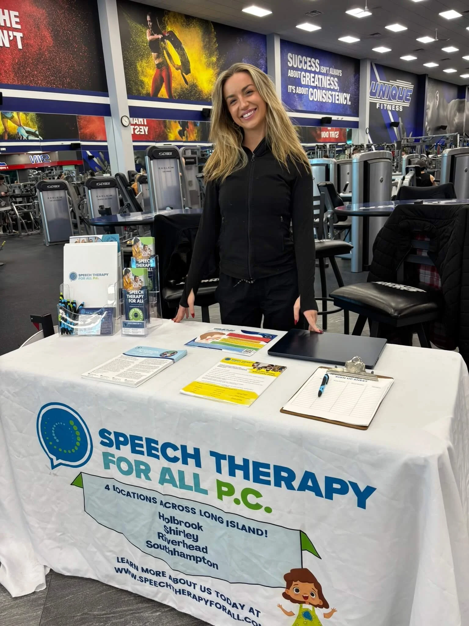A woman standing behind a table at a Speech Therapy event inside a gym. The table displays pamphlets and a sign promoting speech therapy services for all ages with locations on Long Island, New York.