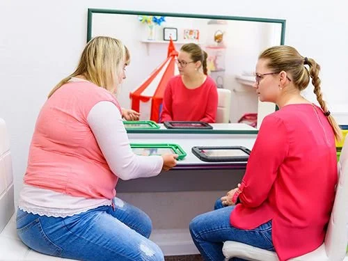 Three women are in a medical or counseling office. One woman, seated behind a glass barrier, is wearing red and glasses. The other two women are seated in front of the barrier, engaging in conversation. One of the women in front is wearing a pink red top and glasses, and the other is in a pink vest and white shirt. There is a collection of trays and paperwork on the table.