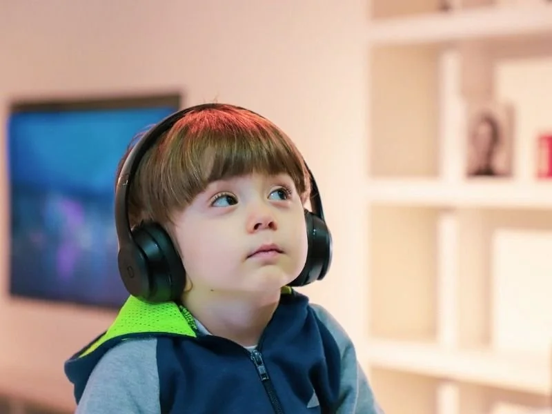 Young boy with brown hair wearing large black headphones and a navy blue hoodie with green accents, sitting in a living room with a TV and bookshelf in the background.