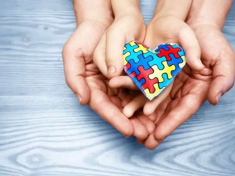 A person's hands gently holding a small stuffed heart with a colorful puzzle pattern on it, against a light wooden surface background.