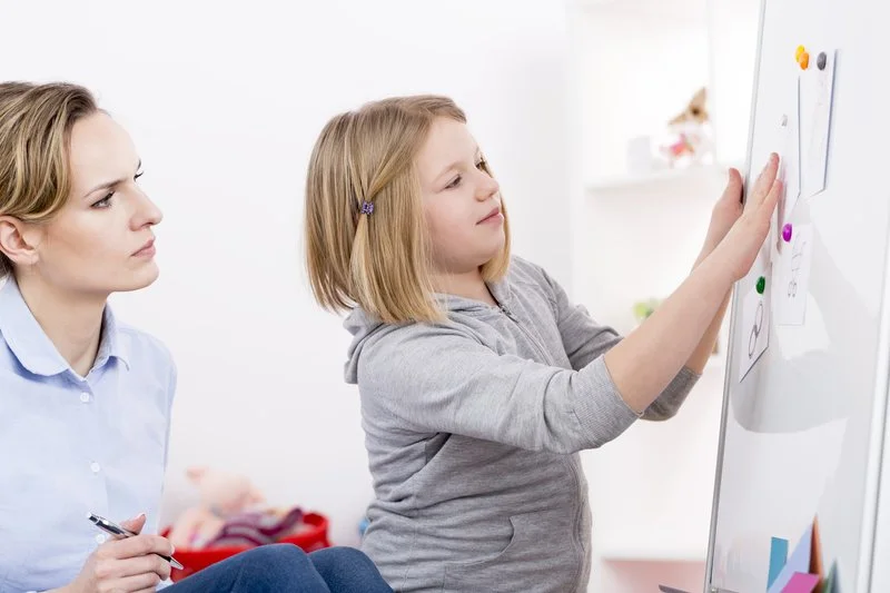Young girl placing a paper with a heart drawing on a whiteboard with magnets, while a woman observes and holds a pen.