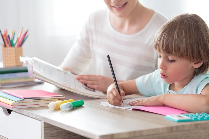 A young boy doing homework at a desk with an adult woman, possibly his mother, helping him. The desk has colorful books, pencils, and a calculator.