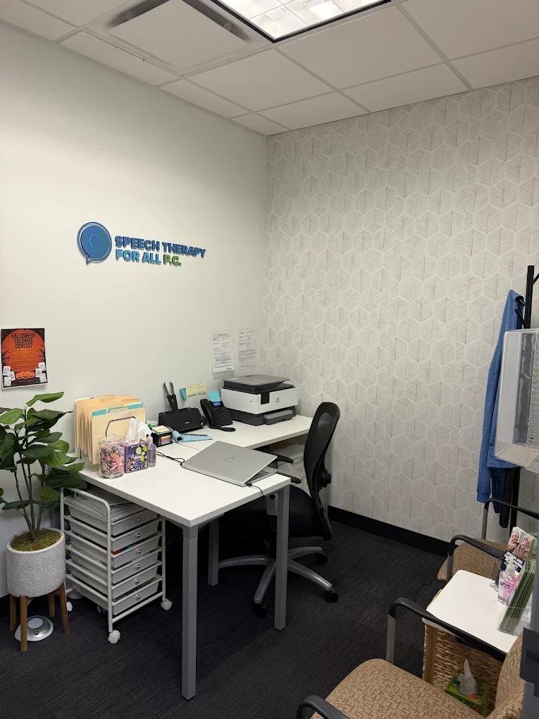Office corner with a white desk, black chair, potted plant, printer, office supplies, and a sign that reads 'Speech Therapy for All PLC' on the wall.