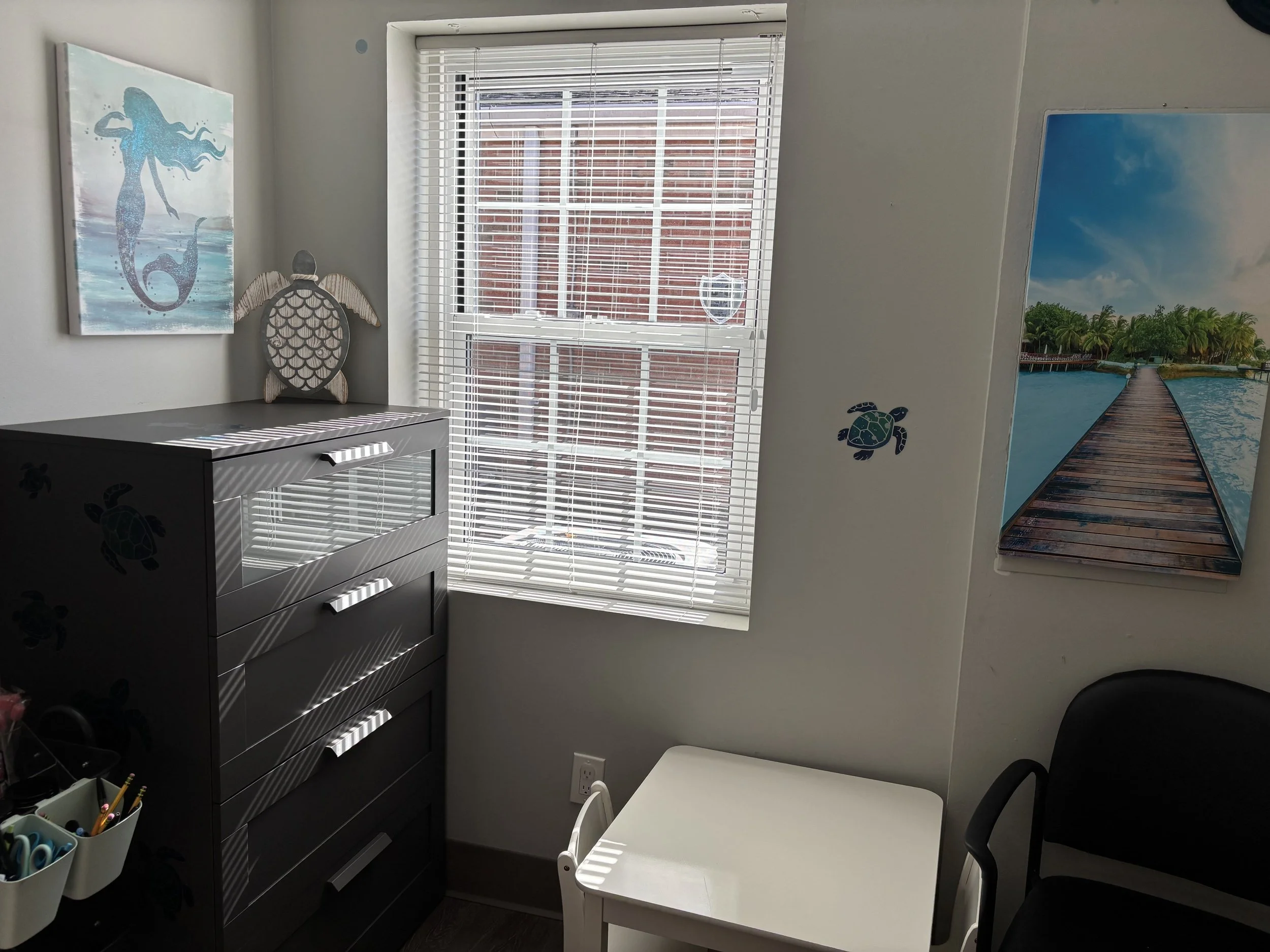 A small room with white walls decorated with ocean-themed artwork and turtle stickers. There is a window with blinds letting in sunlight. A gray dresser with three drawers is against the wall, topped with a wooden turtle decoration. Next to the dress