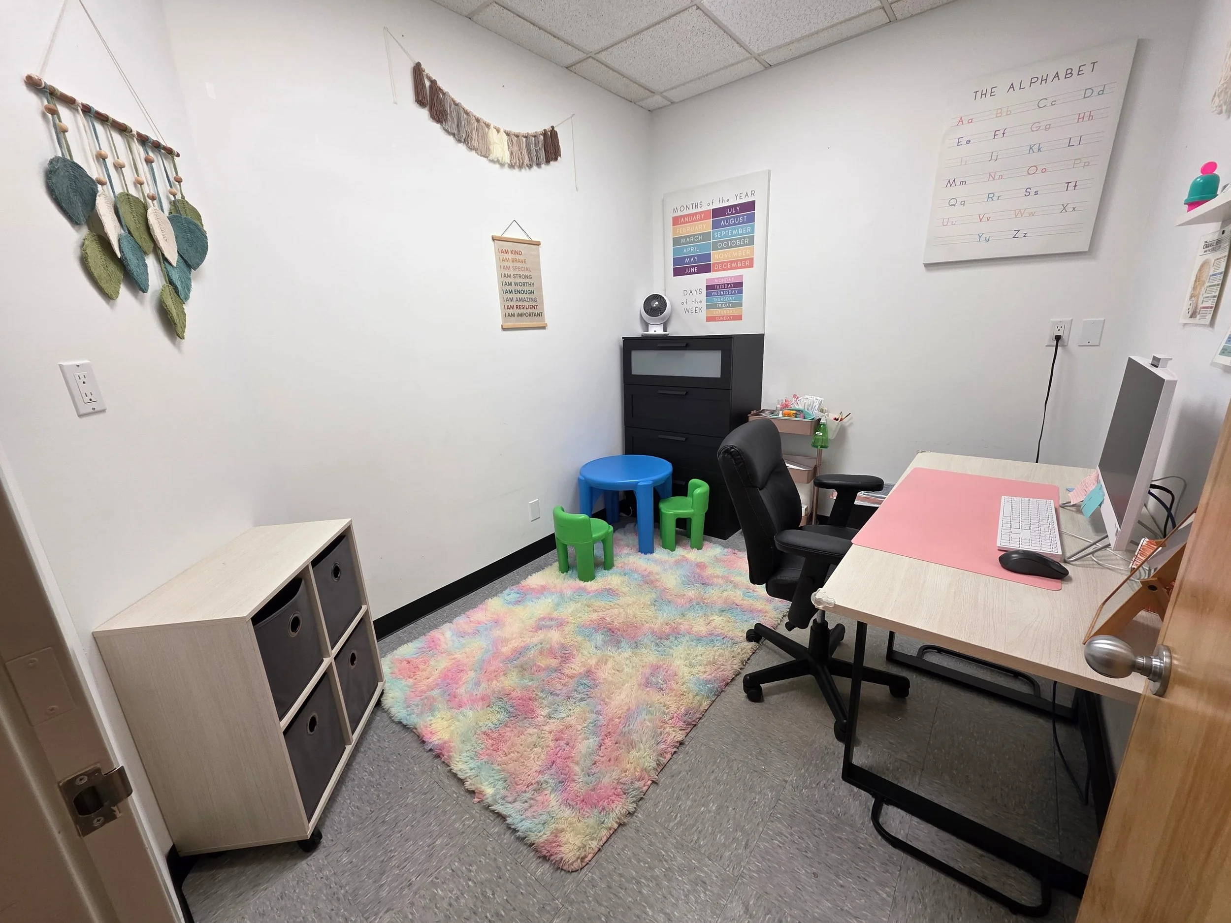 A small, neatly organized classroom or therapy room with a fluffy multicolored rug, a desk with a computer and a pink mouse pad, a black swivel chair, a black cabinet with a fan on top, a small table with colorful chairs, and various educational post