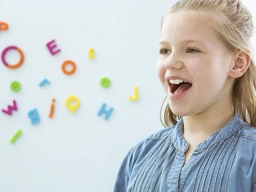 A young girl with blonde hair smiling and looking to the side, with colorful magnetic letters on a white background.
