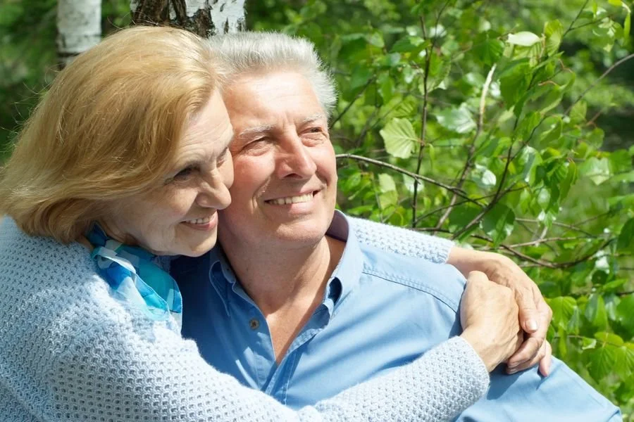 An elderly couple hugging outdoors among green leaves.