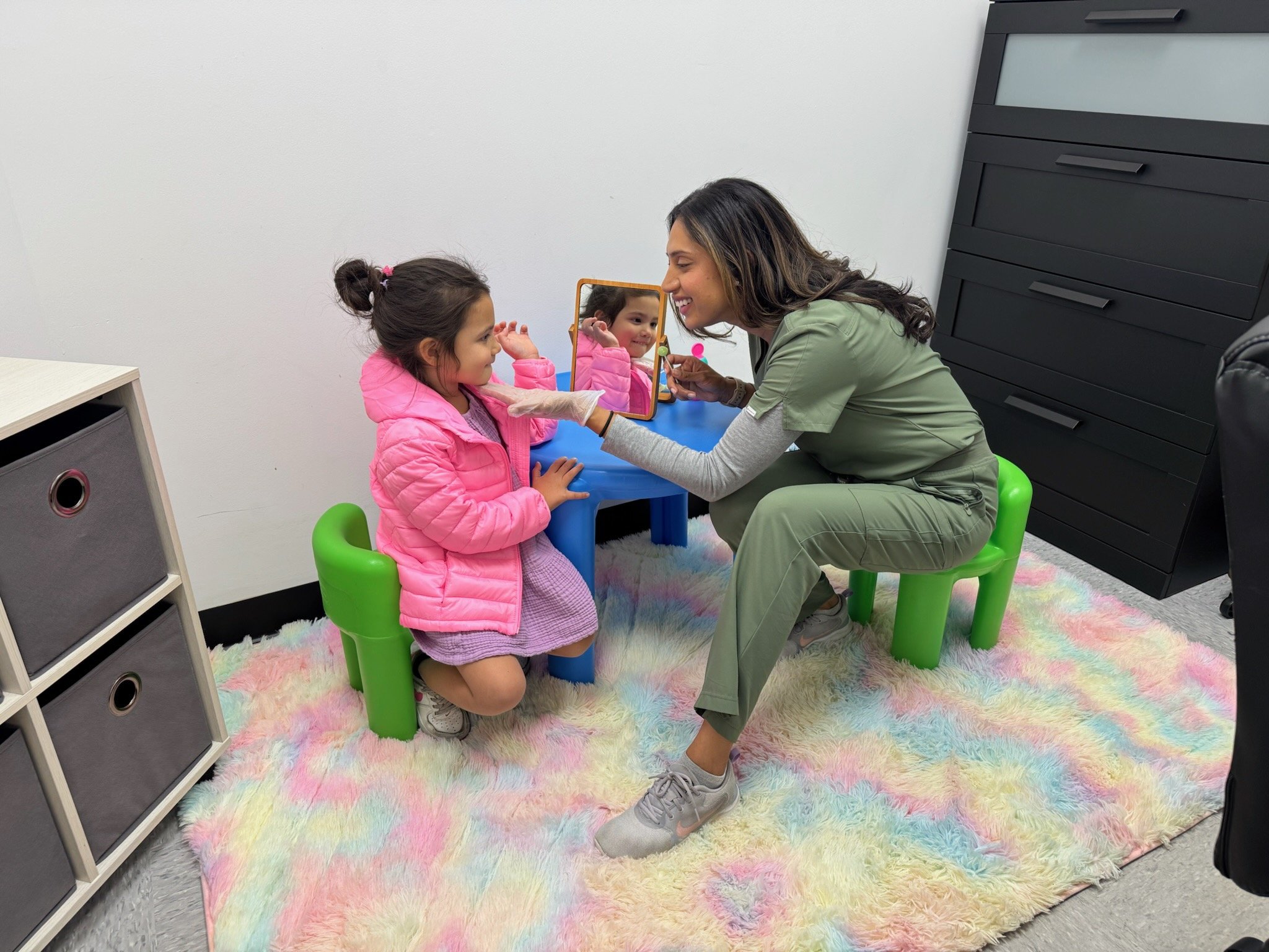 A woman in a green medical uniform and gloves is smiling as she applies lipstick to a young girl in a pink jacket at a small blue vanity table. The girl, who has dark hair in a bun, is sitting on a green chair, looking into a mirror. They are in a room with a colorful fluffy rug, a white wall, a black storage cabinet, and a cubby with fabric drawers.