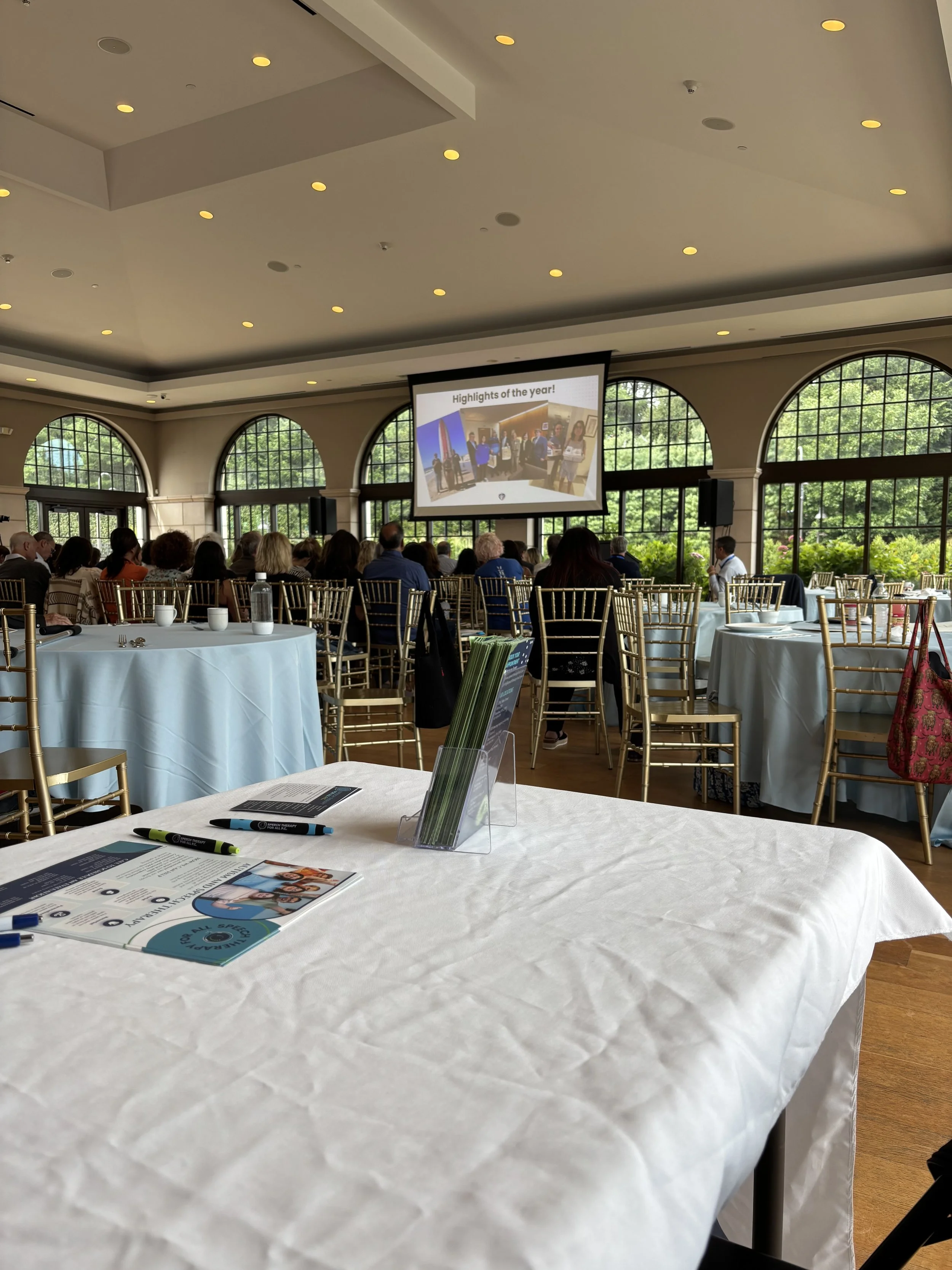Conference room with attendees watching a presentation on a large screen, tables with chairs, and large windows with greenery outside.