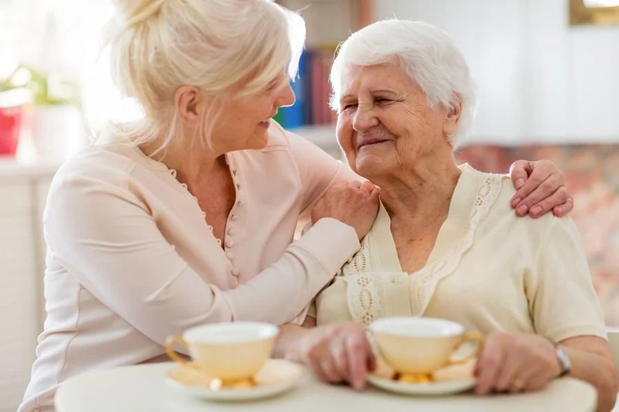 An elderly woman and a middle-aged woman sharing a moment at a table with tea cups, smiling at each other.