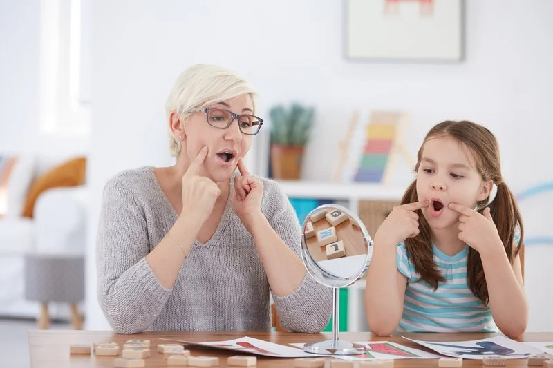A woman and girl making funny faces at a desk with ruler, papers, and a mirror; in a bright room with a bookshelf and colorful decor.