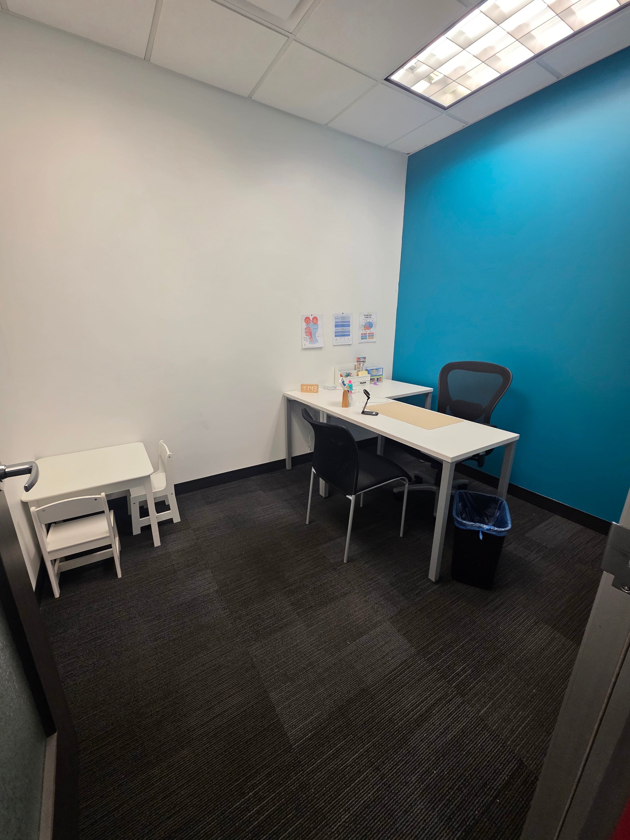 An empty medical consultation room with a white and blue wall, a white desk with chairs, medical supplies, and a trash can.