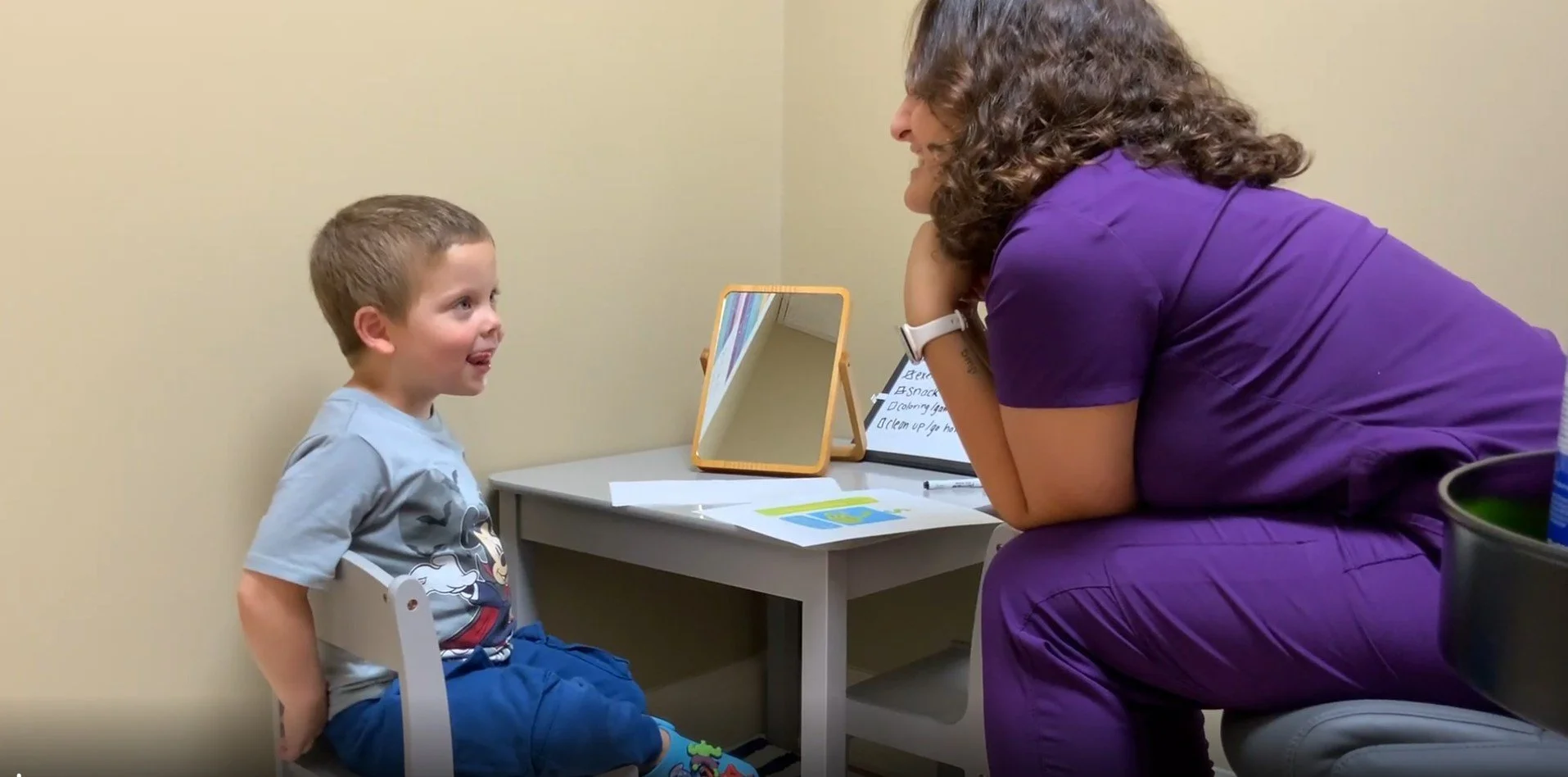 A young boy sitting on a small chair talking to a woman, likely a doctor or therapist, in a consultation room.