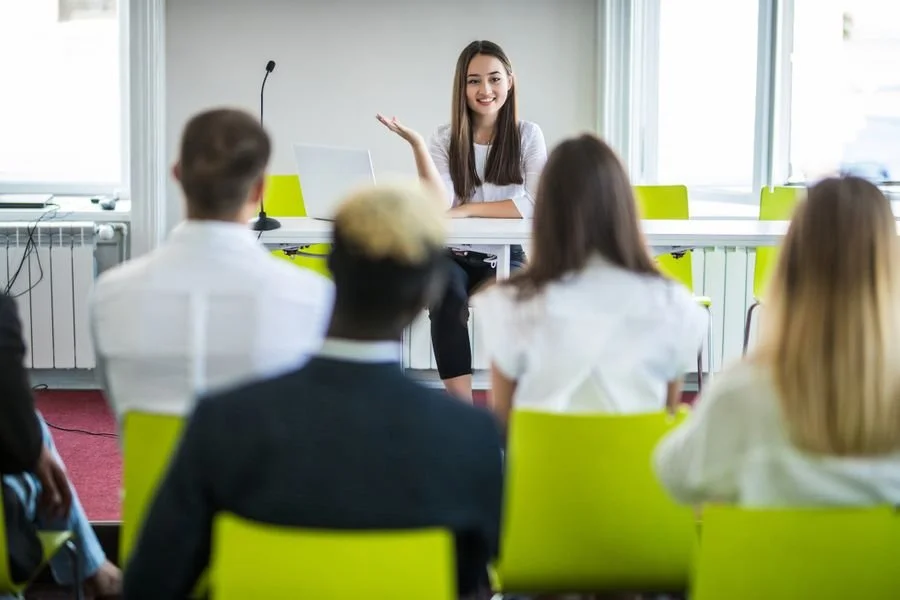 A woman giving a presentation or lecture to a group of people in a conference room with large windows.