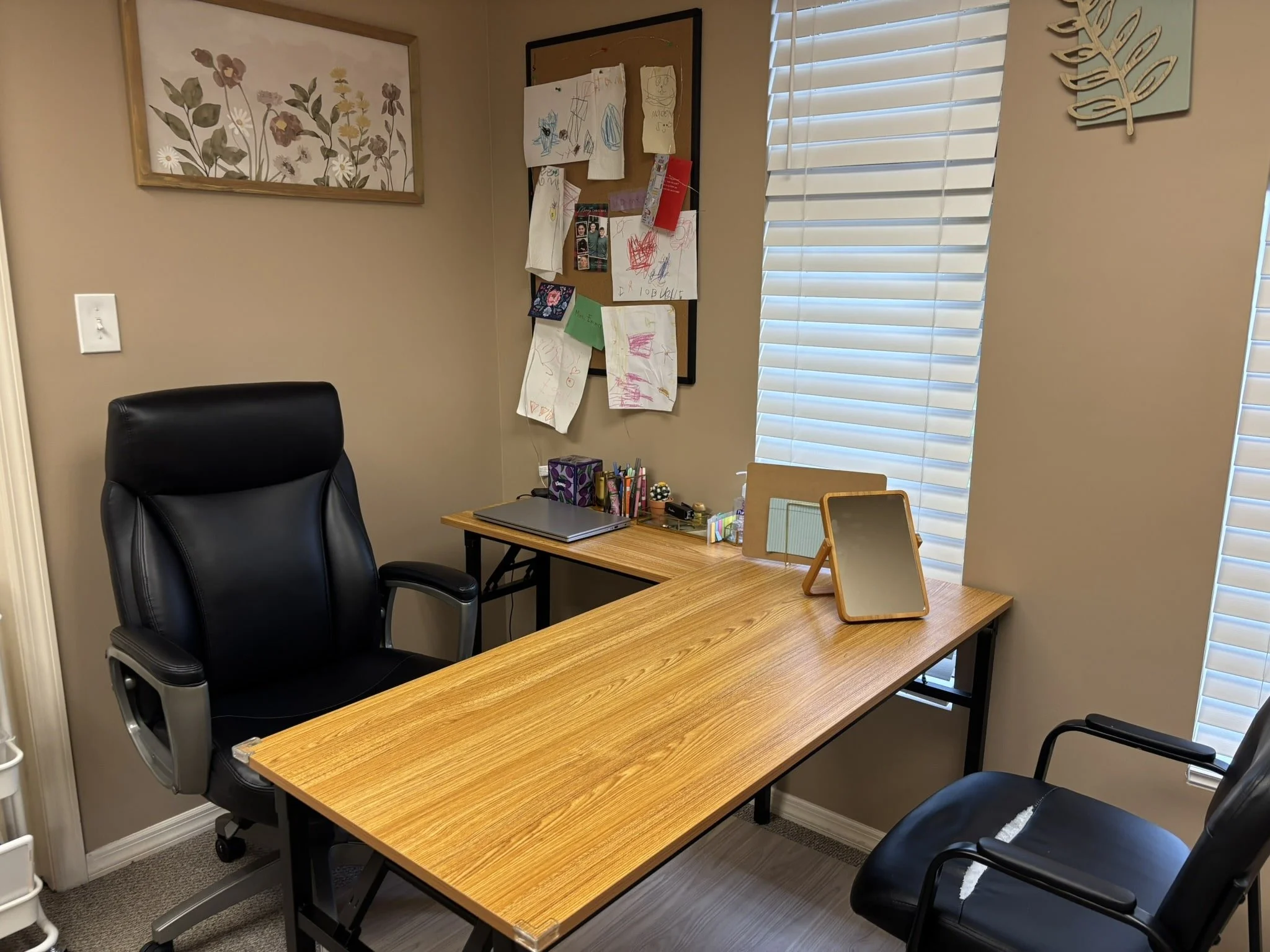 An office space with two black chairs, a wooden desk, and a wall decorated with artwork, a bulletin board with children’s drawings, and window blinds.