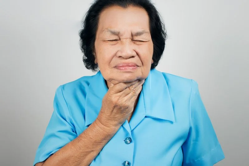 Older woman in a blue shirt holding her neck, appearing to have throat pain or discomfort.