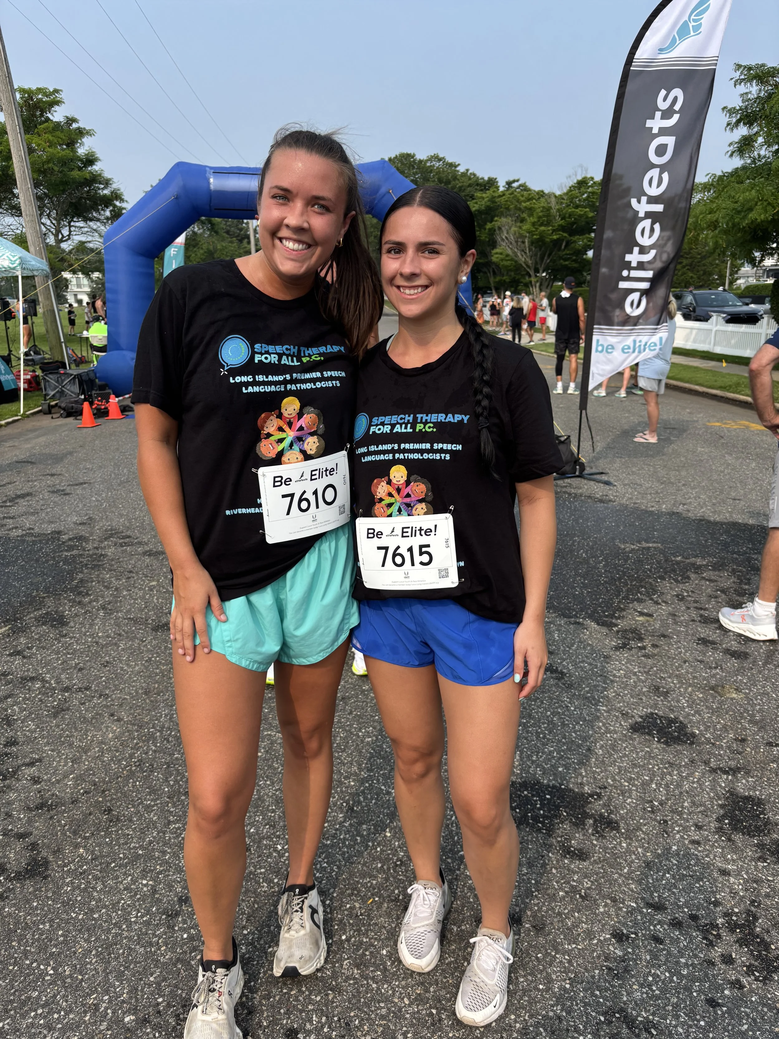 Two women wearing black t-shirts and athletic shorts, standing outdoors after a race, smiling at the camera. They have race bibs with numbers 7610 and 7615. In the background, there are other people, trees, and event banners, including a prominent black and white banner that says 'elite' and a blue inflatable arch.