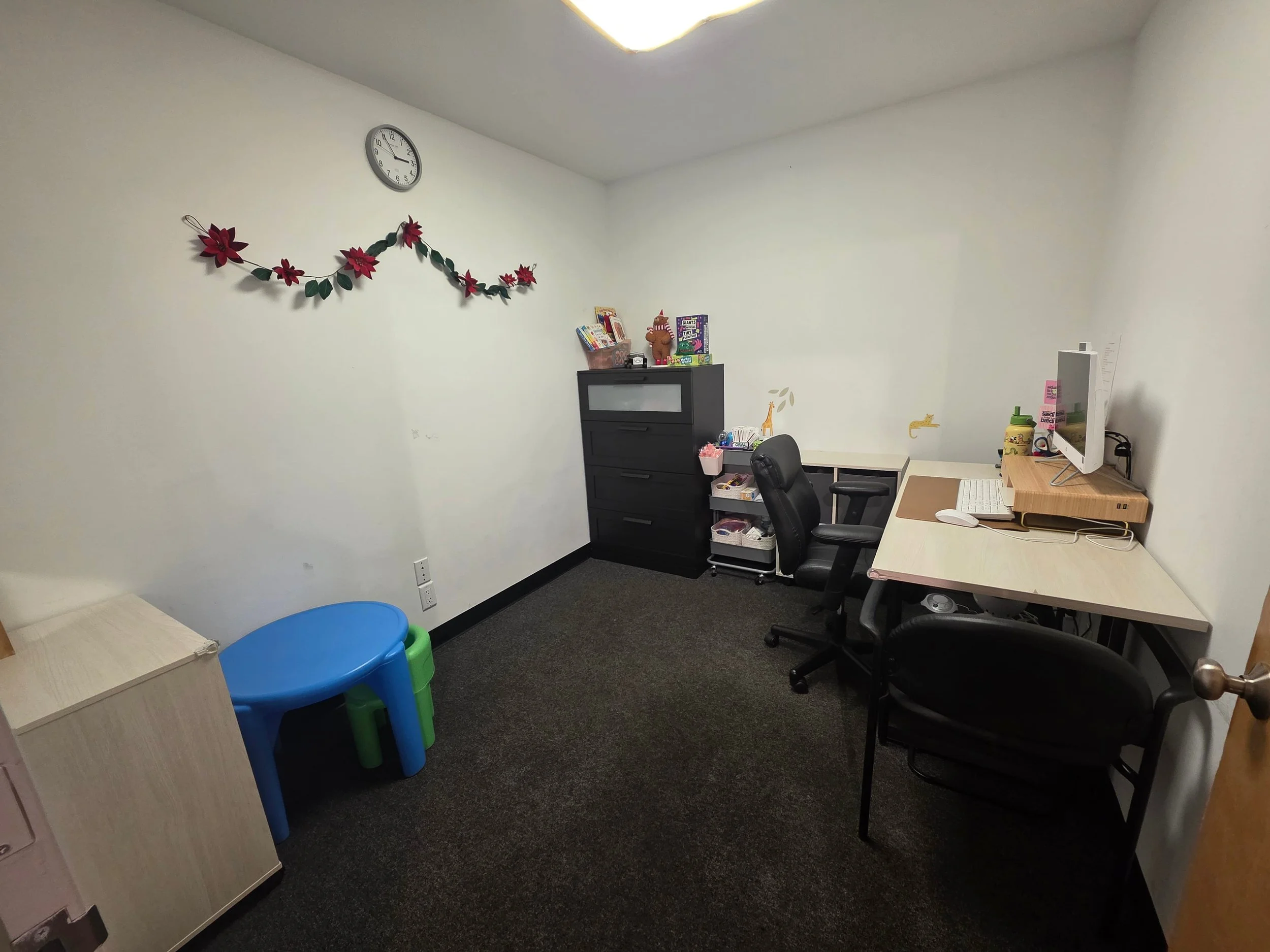 A small office room with white walls, black carpet, and a simple arrangement of furniture, including a white desk with a computer and a black office chair, a black drawer unit, a small blue and green plastic table, a wooden cabinet, and decorative Ch