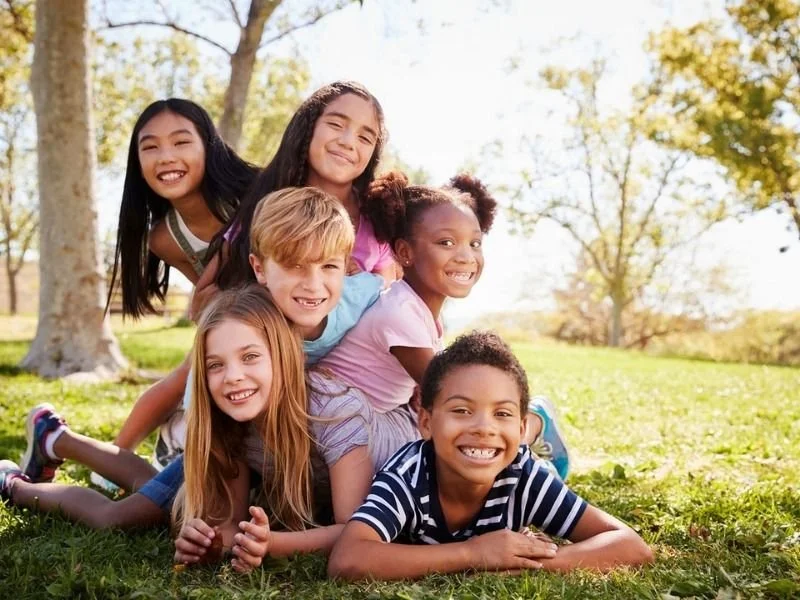 Group of six children, five girls and one boy, smiling and lying on grass in a park with trees in the background.