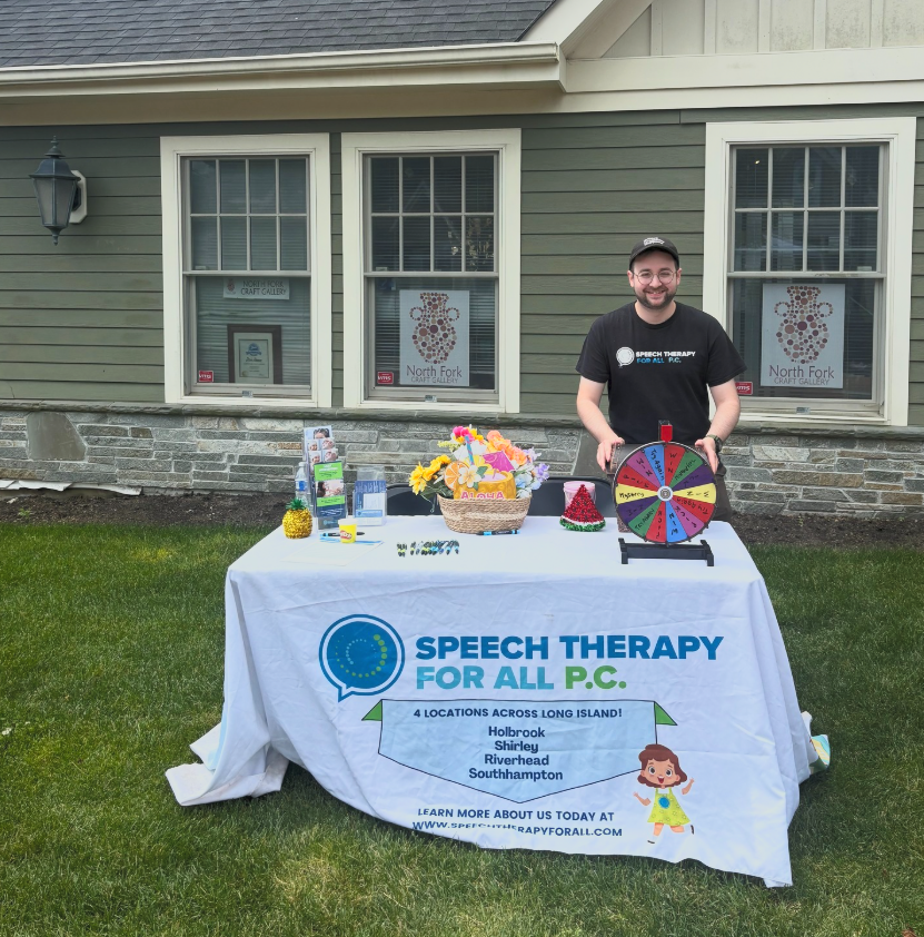Young man standing behind a table setup outdoors with a green house in the background. The table has a white tablecloth with a speech therapy organization’s logo and information, decorated with colorful flowers, a small pineapple, a basket with flowers, a red cone-shaped object, and a colorful spinning wheel.