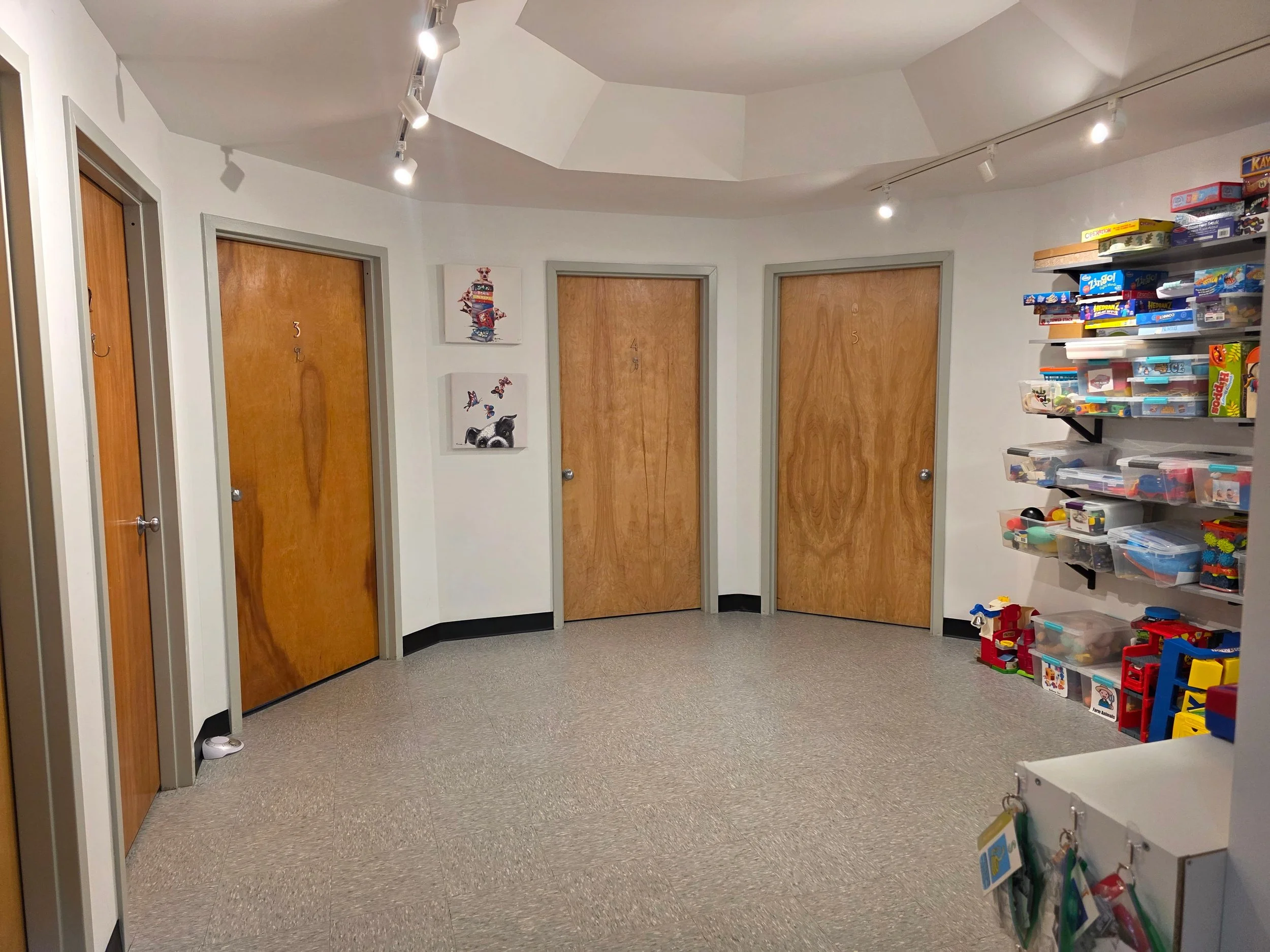 A small, brightly lit room with three closed wooden doors, each labeled with a number: 3, 4, and 5. The room has white walls, a textured beige floor, and a white ceiling with track lighting. There are colorful toys and storage containers on a wall-mo
