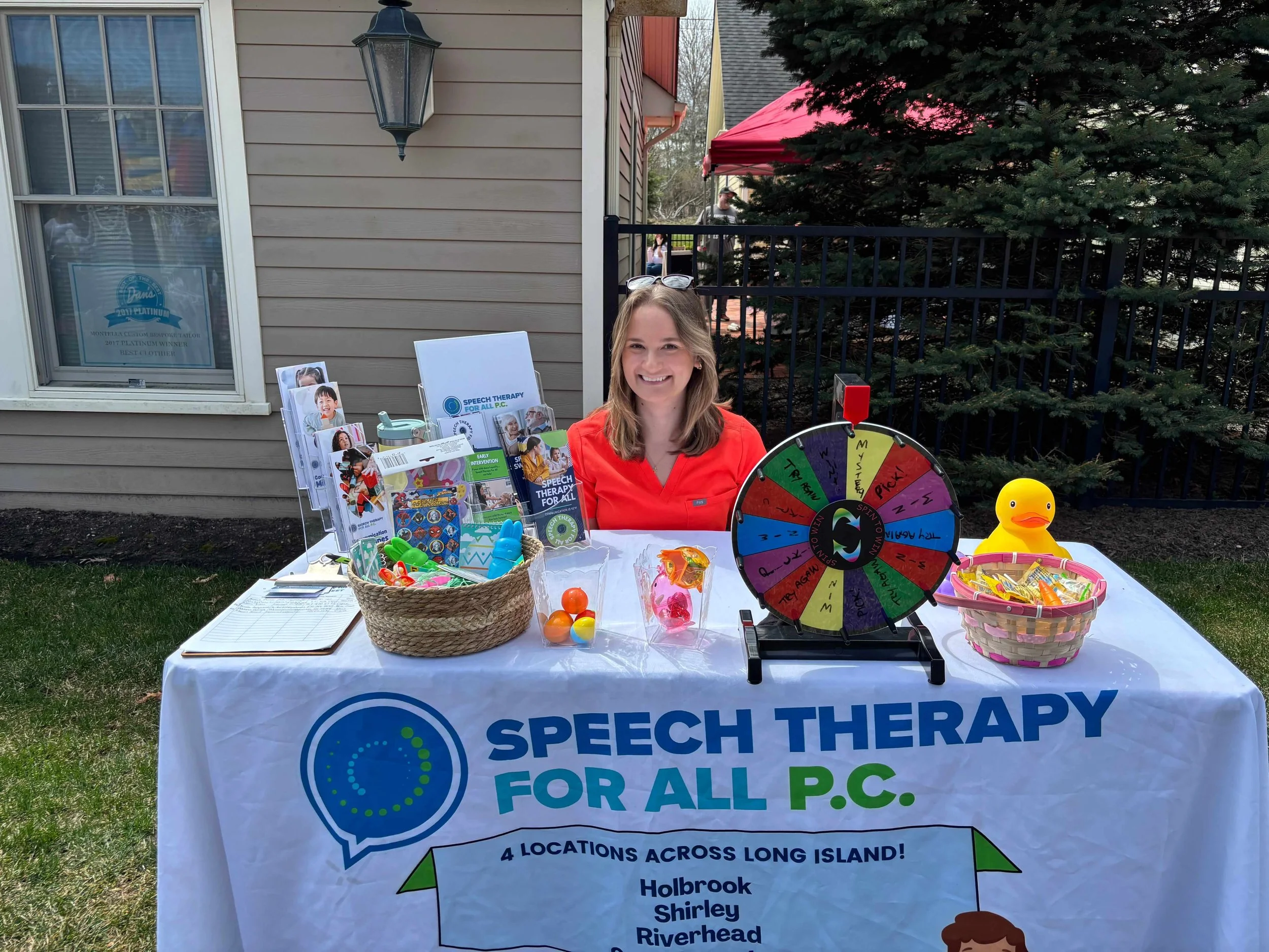 A woman sitting at a table outdoors, promoting speech therapy services. The table has a sign reading "Speech Therapy for All P.C." with four locations listed. On the table are a spinning wheel, toys, informational pamphlets, a basket of items, and a rubber duck. The woman is smiling and wearing a red shirt, with a house and trees in the background.