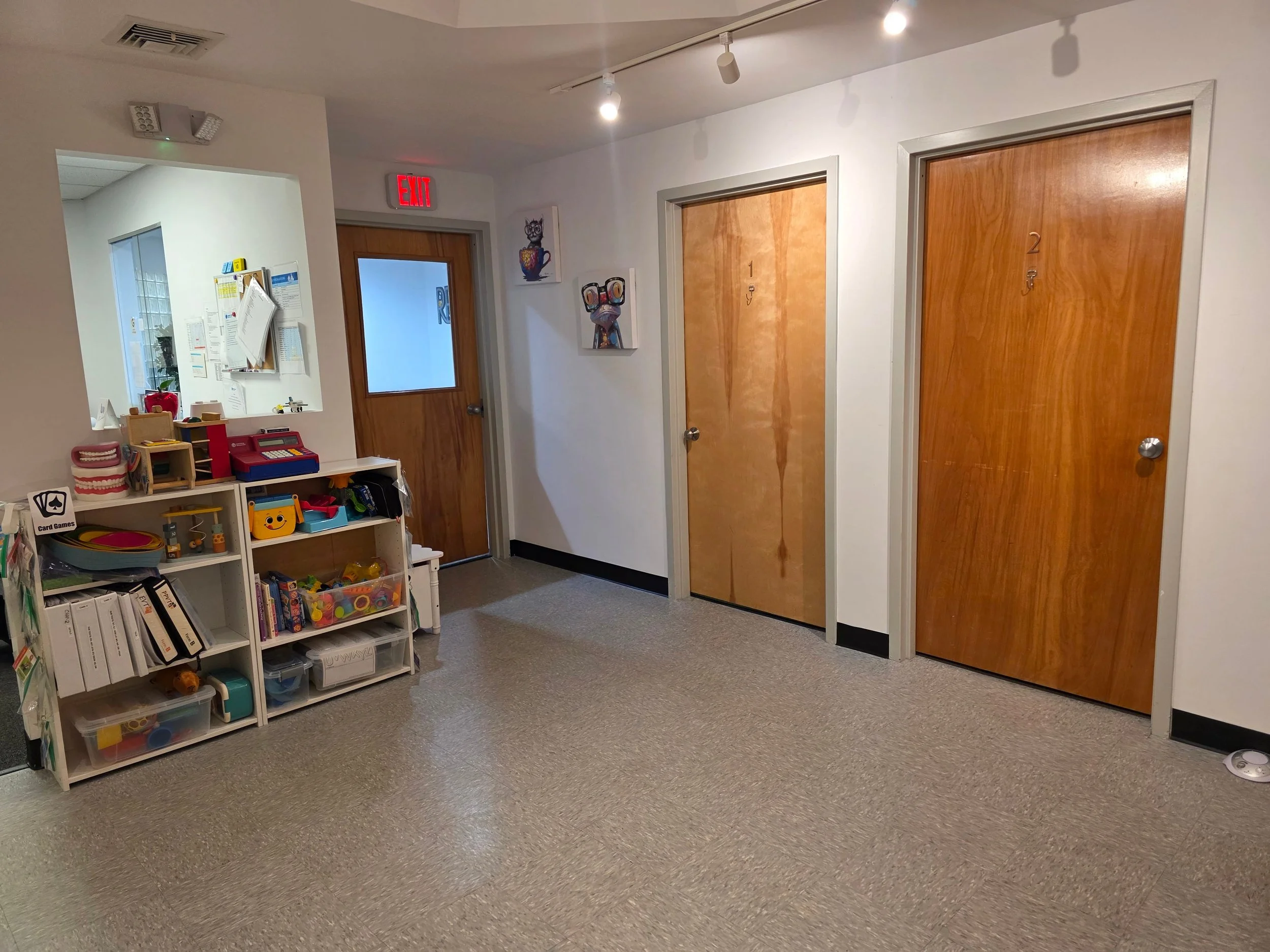 Interior of a children's play area or daycare with cubbies filled with toys and books, two closed wooden doors, colorful artwork on the wall, and an office door with a window.