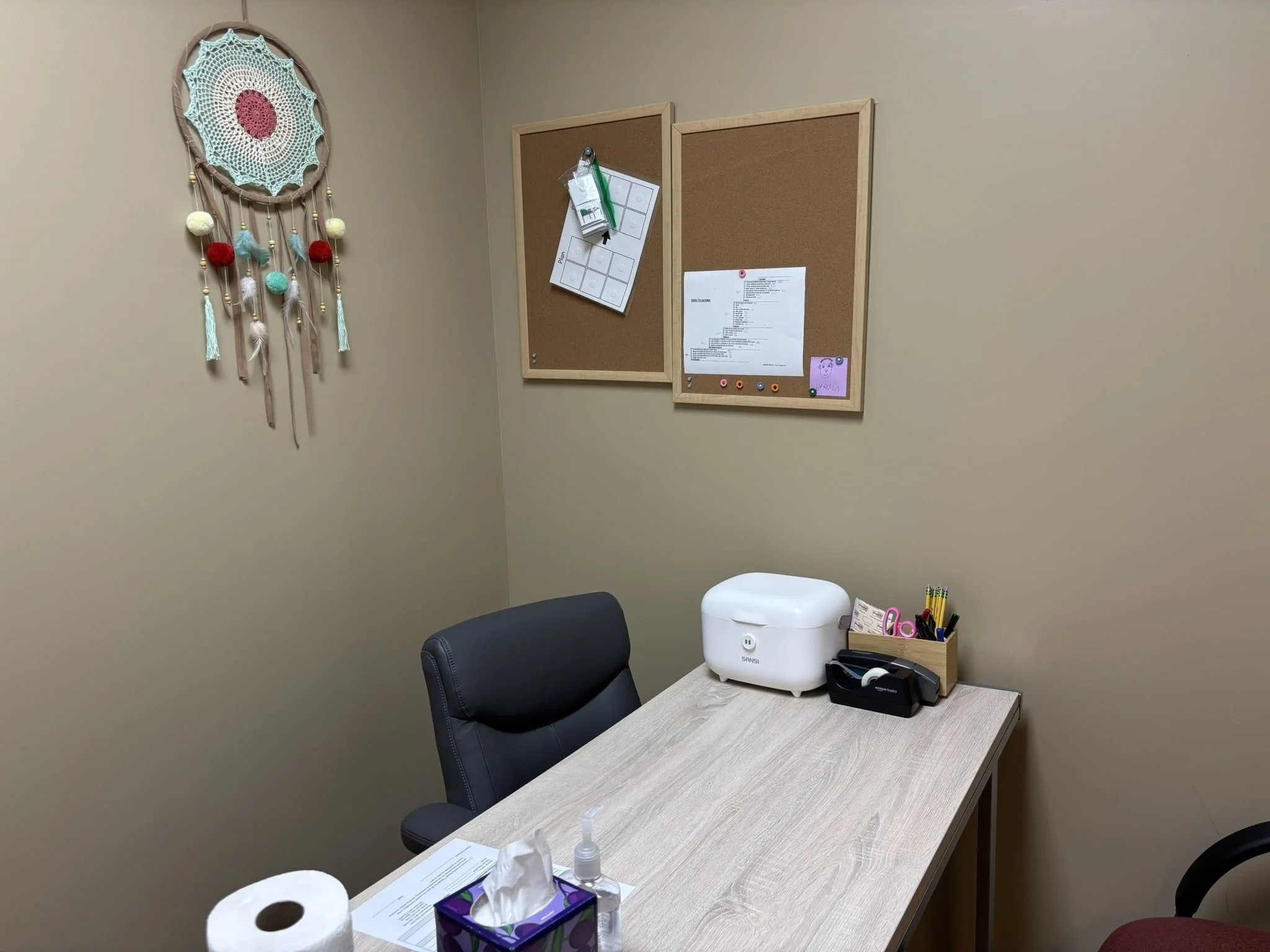 Office or consultation room with a desk, black swivel chair, tissue box, hand sanitizer, toilet paper on the desk; corkboards on the wall, one with papers and a small purple drawing. Decorated wall hanging resembling a dreamcatcher with colorful ball