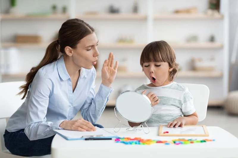 A woman, possibly a therapist or teacher, sits across from a young boy at a white table. The woman is holding up her hand in a stop gesture, and the boy is looking into a small mirror with an surprised or amused expression. The table has colorful alphabet letters and a tablet. The background shows a light, airy room with shelves and various items.