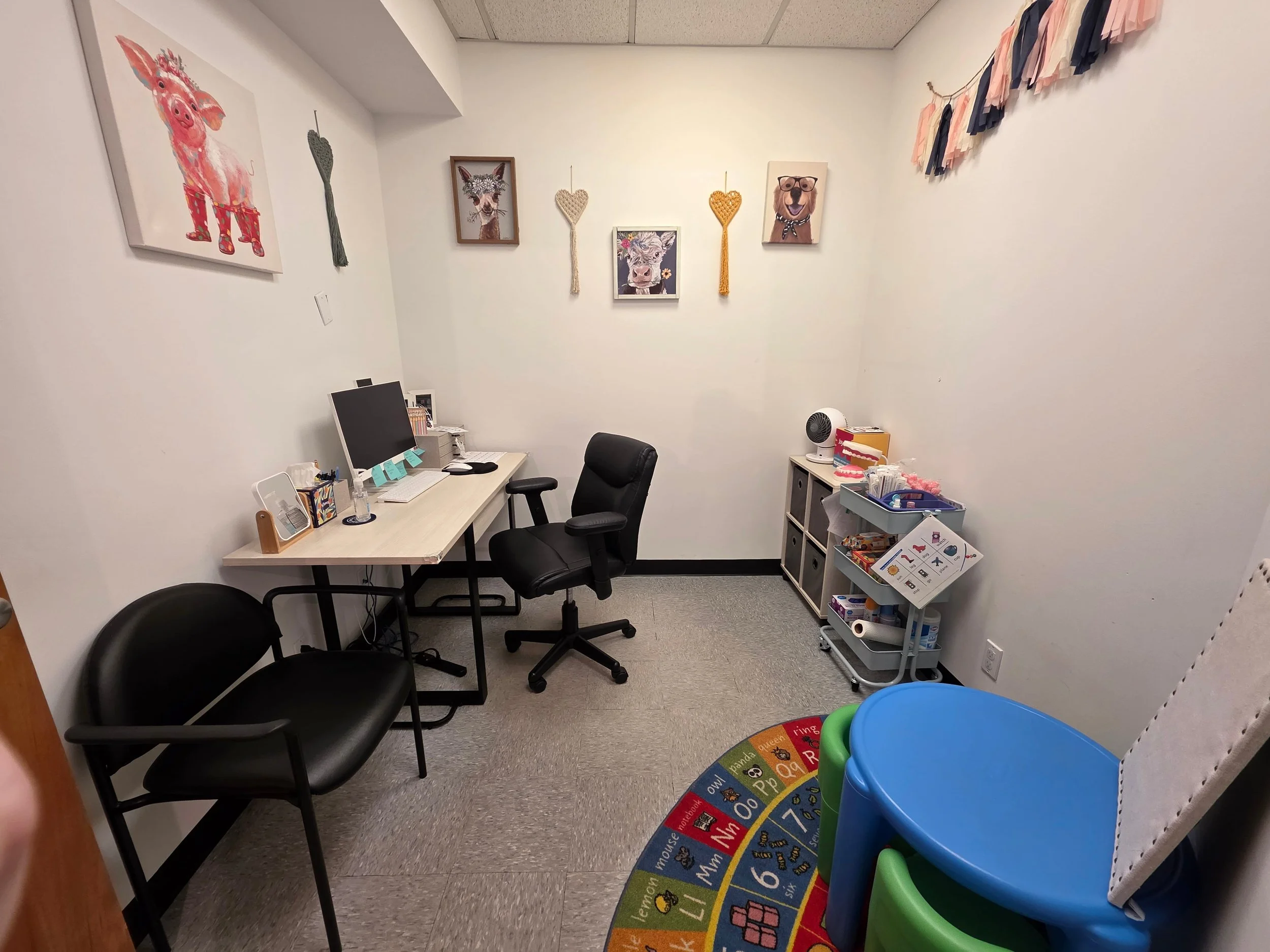 An office and craft room with a white desk, black office chair, two black visitor chairs, a colorful children's play rug, and wall art including animal portraits and heart decorations.