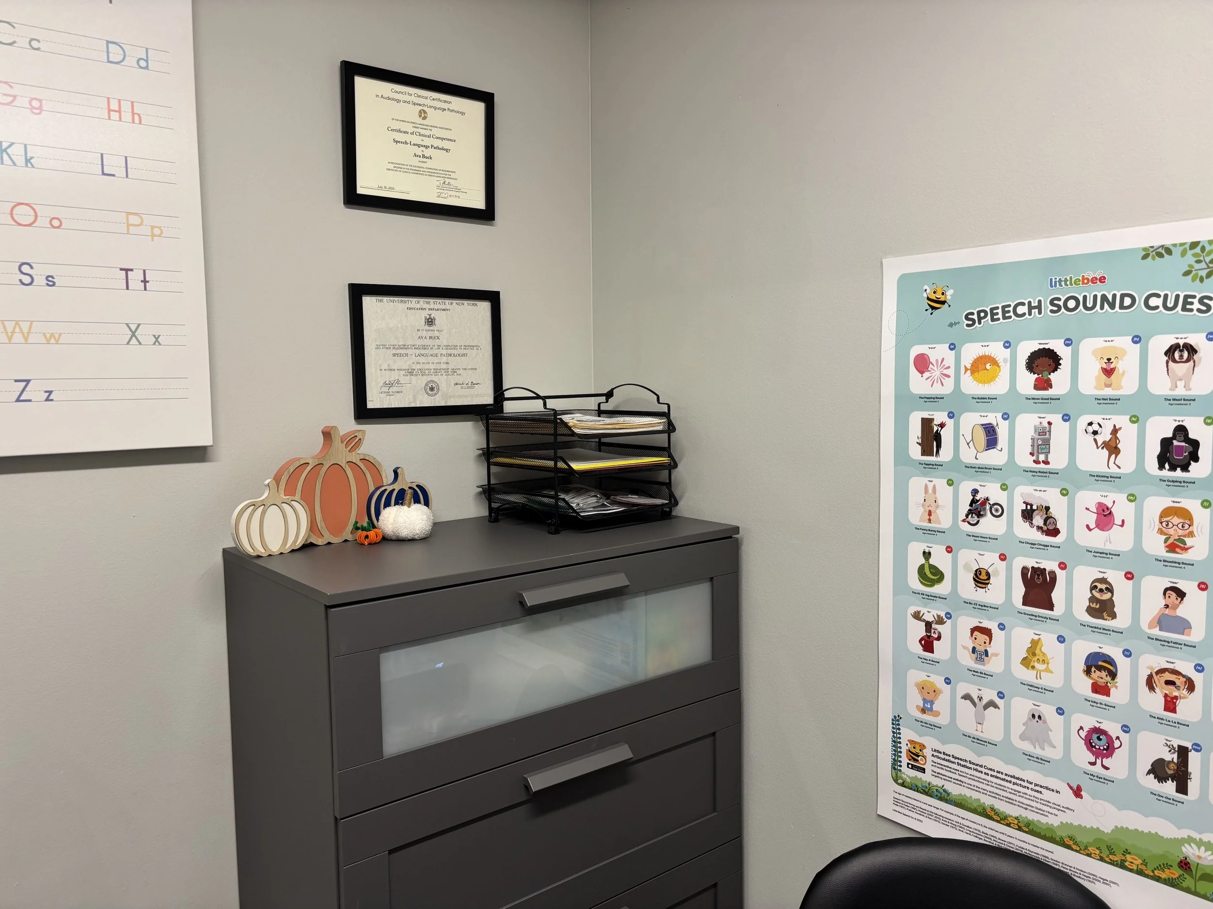 A gray cabinet with three drawers and a glass top, decorated with fall-themed pumpkins and a small pumpkin figurine, situated in an office with framed certificates on the wall and a colorful speech sound cues poster to the right.