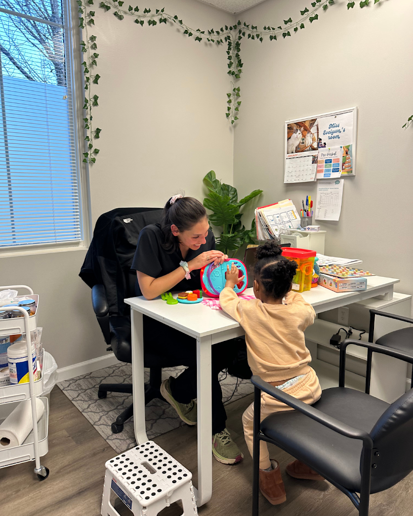 A woman and a young girl playing with toys at a desk in a medical or therapy office, with a window, potted plant, and a bulletin board in the background.