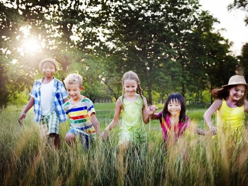 Five children holding hands and running through a grassy field in a park during sunset, smiling and enjoying outdoor activity.