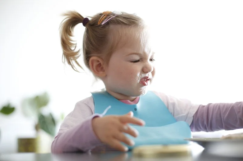 A young girl with a messy face, wearing a bib, scrunching up her face while eating or playing with food at a table.