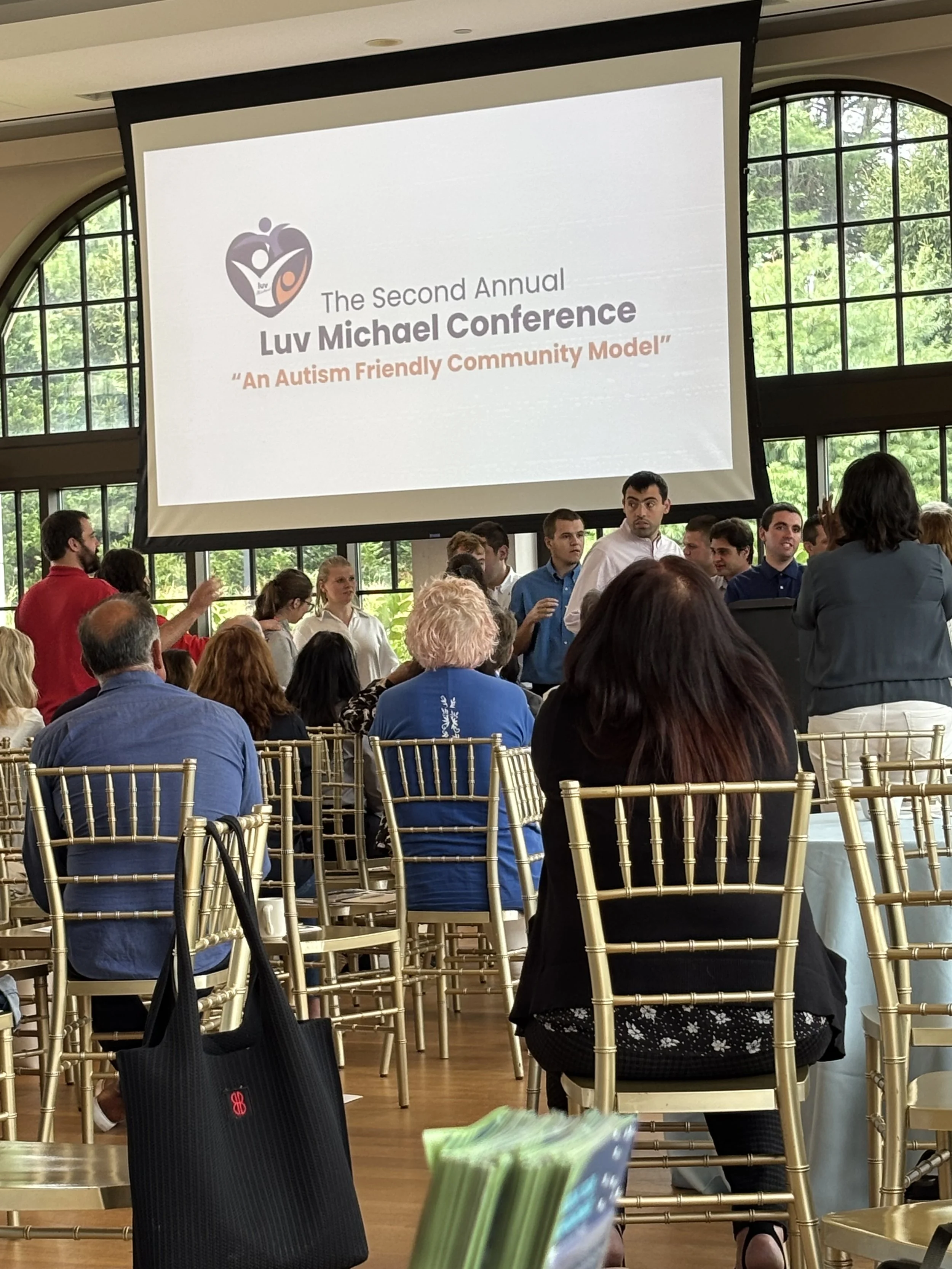 A conference room filled with attendees seated on gold-colored chairs, facing a large screen displaying a presentation slide titled 'The Second Annual Luv Michael Conference 'An Autism Friendly Community Model'' with a logo above.