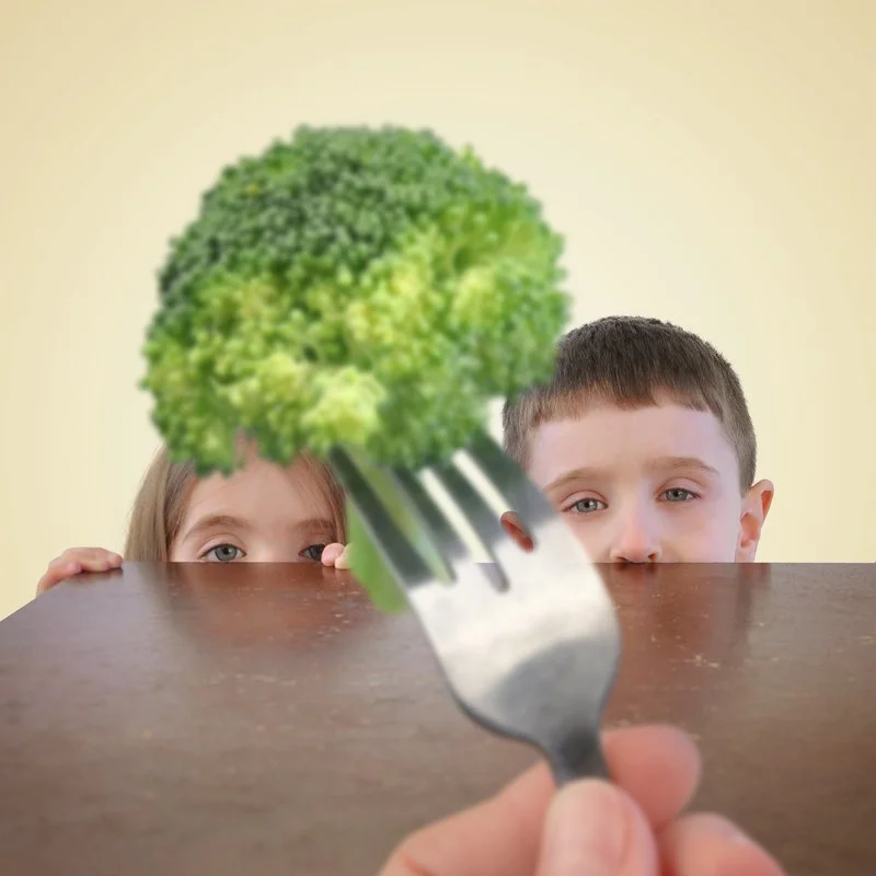 Two children peeking over a table with a fork holding a piece of broccoli in front of the camera.