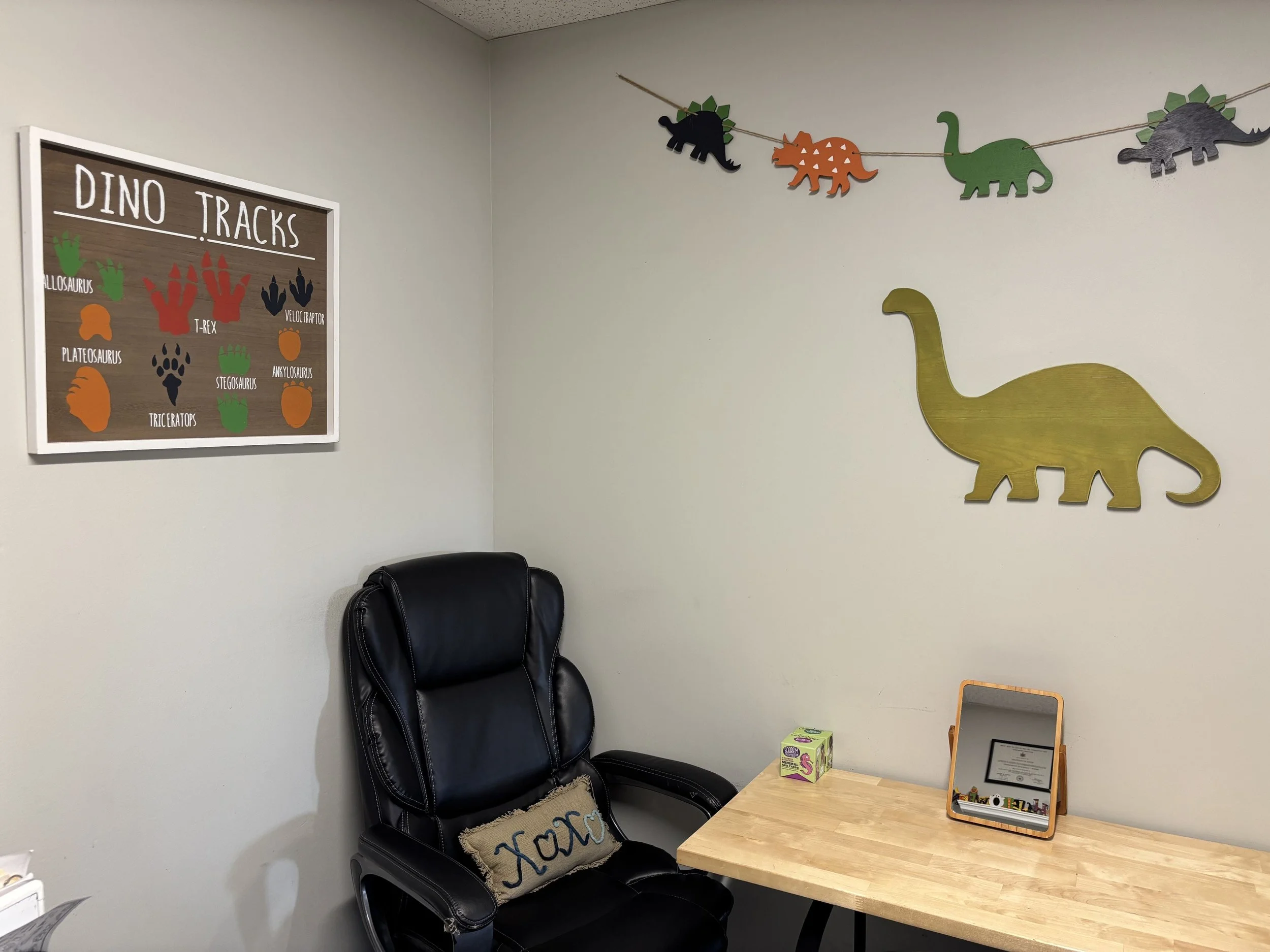 A corner of a child's room with dinosaur decor. There is a cream wall with a large gold dinosaur cutout and a string of colorful dinosaur paper cutouts. A black chair with a cushion labeled 'Xom' is next to a wooden desk with a small mirror and a box