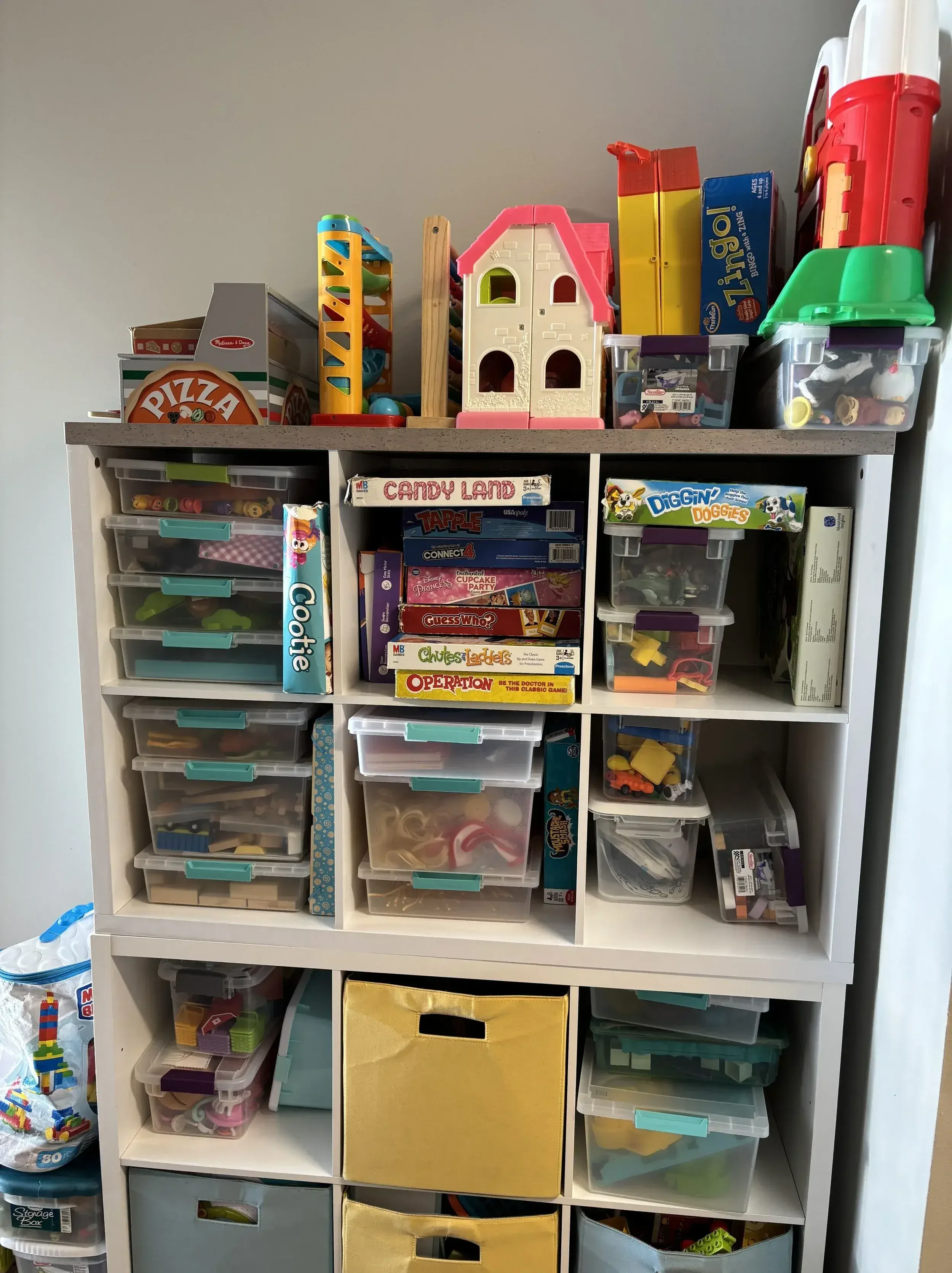 Toy storage unit with various plastic bins containing toys and games, including board games like Candyland, Connect 4, Guess Who, and Operation, and building blocks on top.