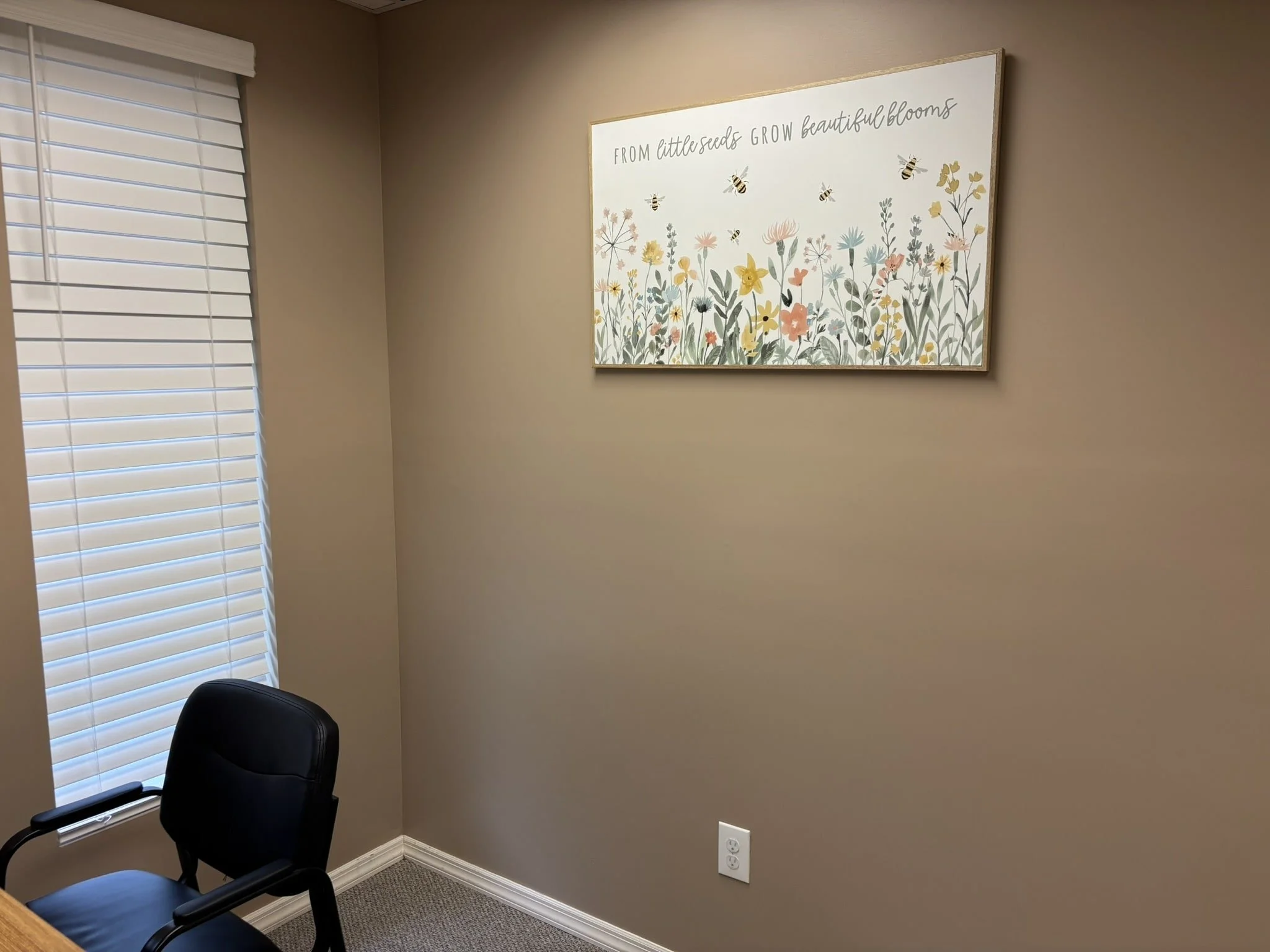 Empty beige office corner with a black chair, a window with blinds, a floral wall art with bees and the quote 'From little seeds grow beautiful blooms'.