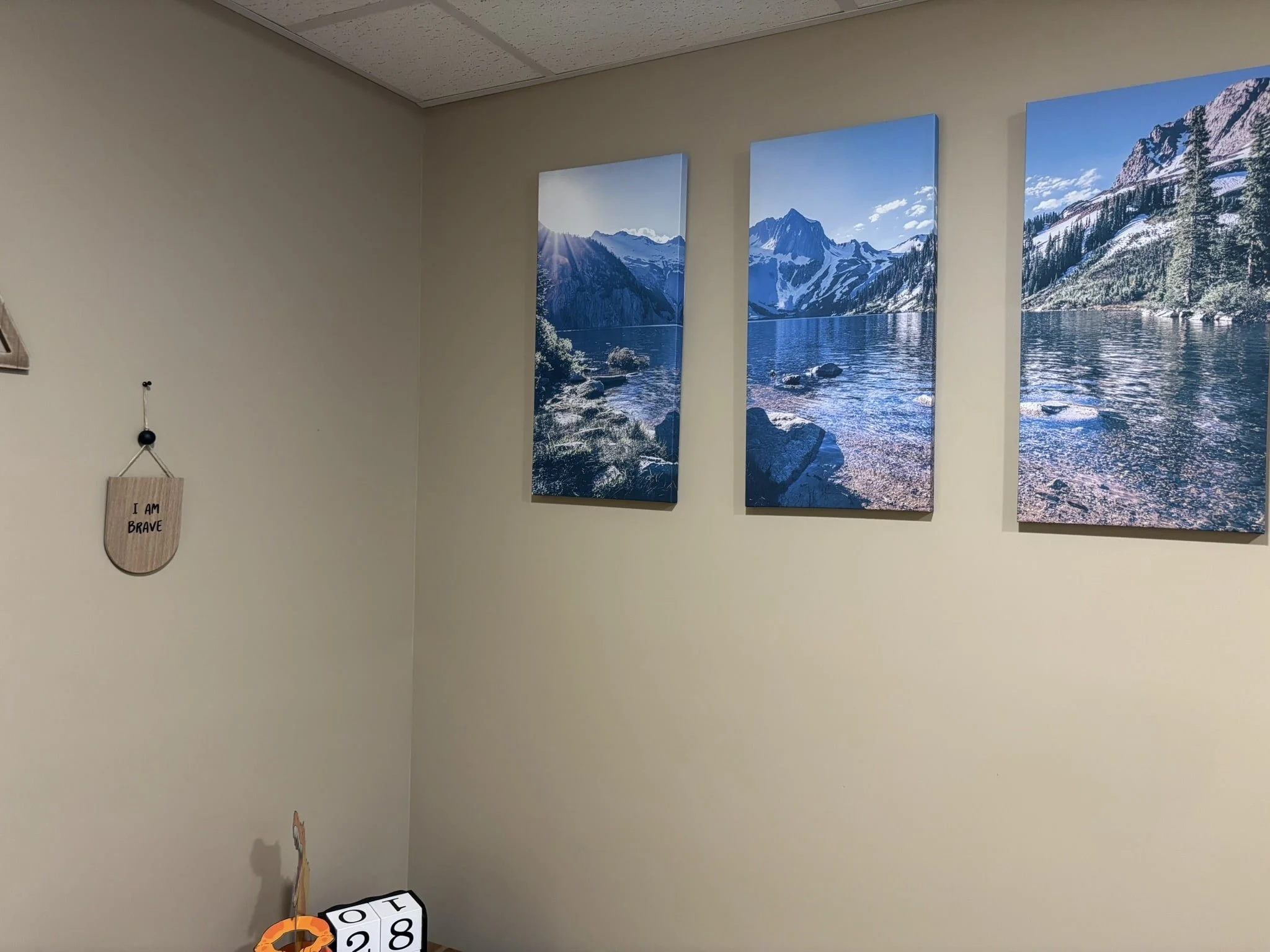 Interior wall with three landscape canvas prints of mountains and a lake, a small wooden sign that says "I AM BRAVE," and a cube calendar showing the date October 28.
